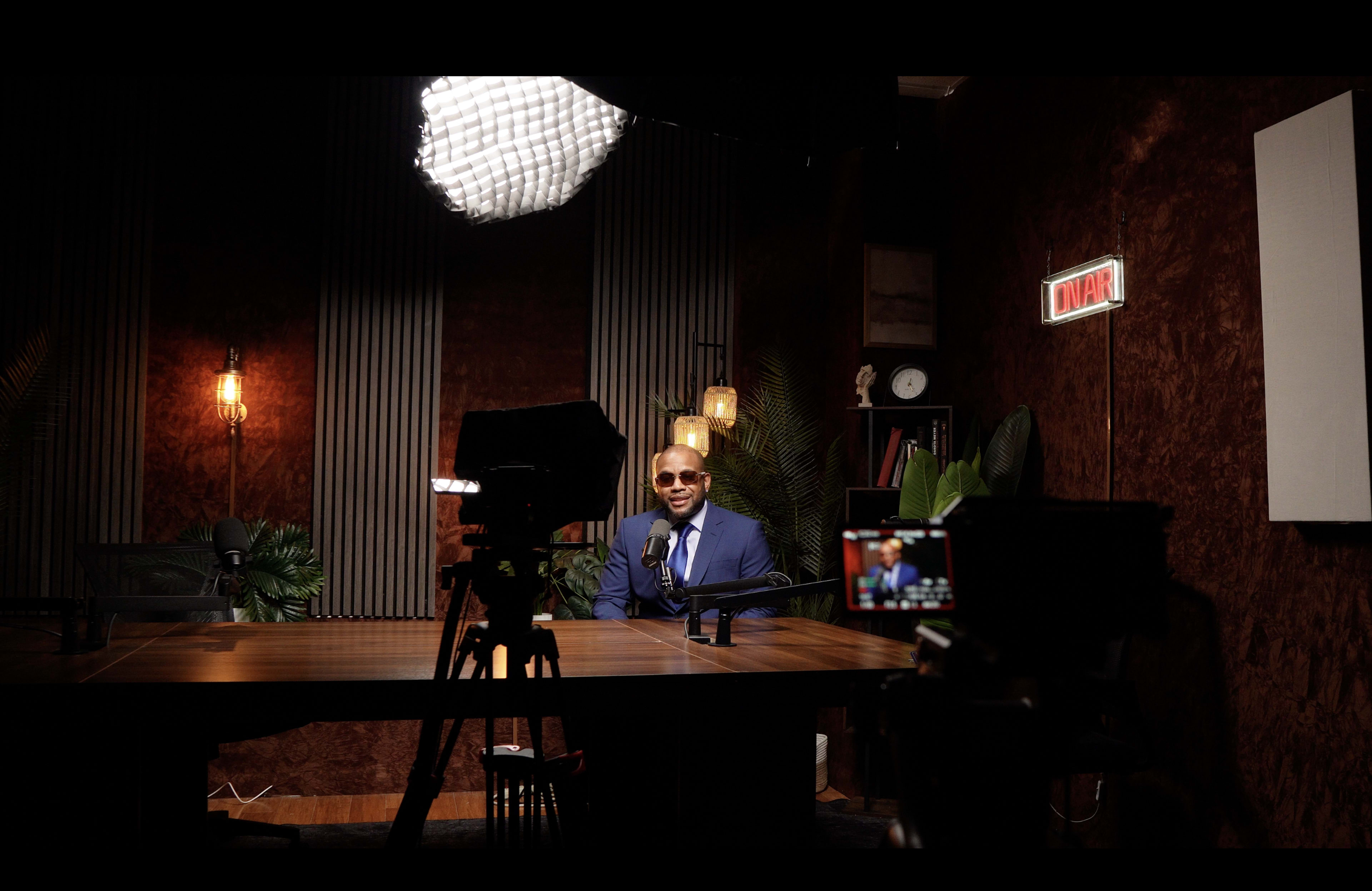 A man in sunglasses sits at a table in a well-lit recording studio with plants and modern decor.