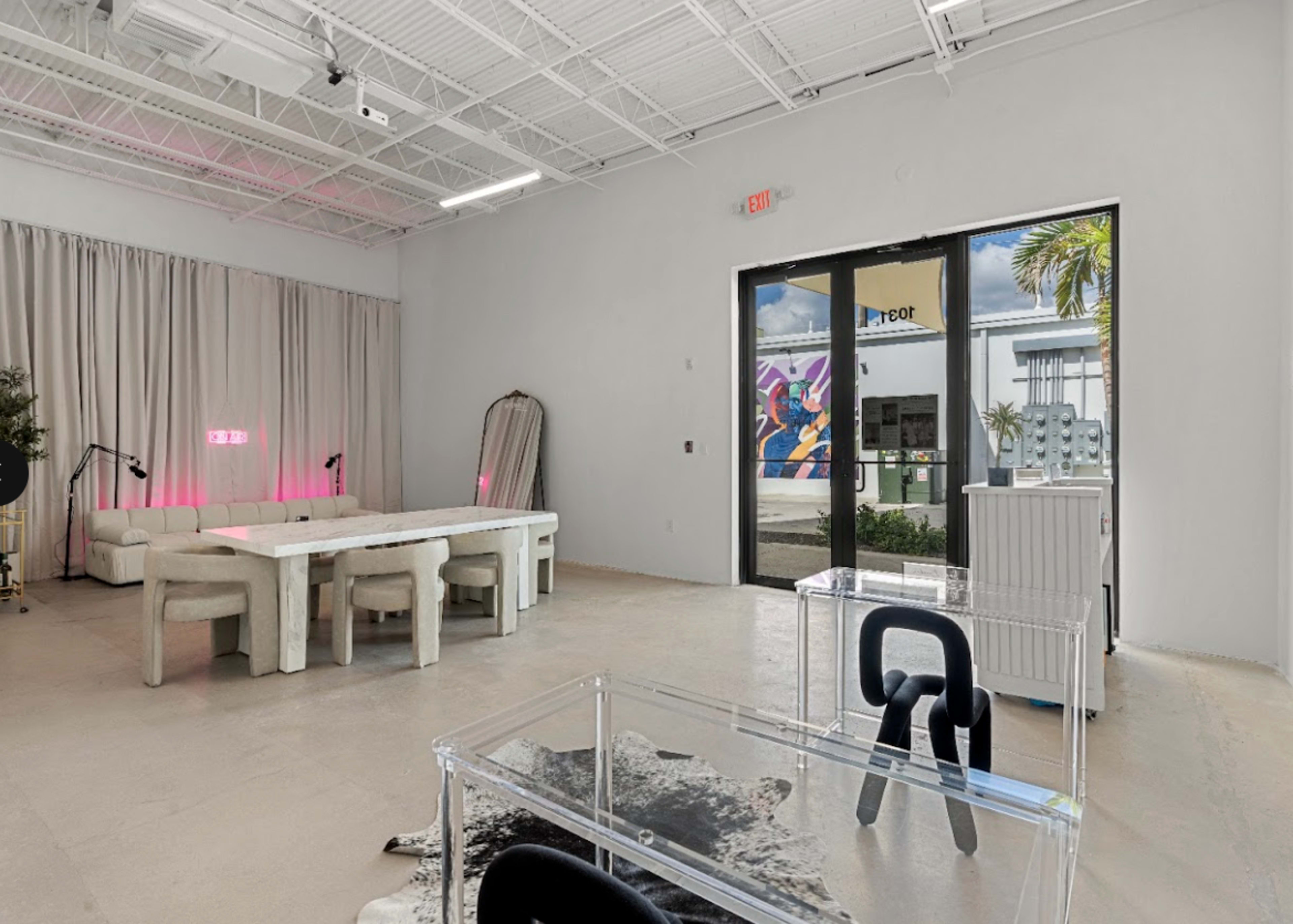 The image shows a spacious modern interior featuring a dining table surrounded by chairs, a large mirror, and an adjacent glass door leading to an outdoor area with murals and greenery.