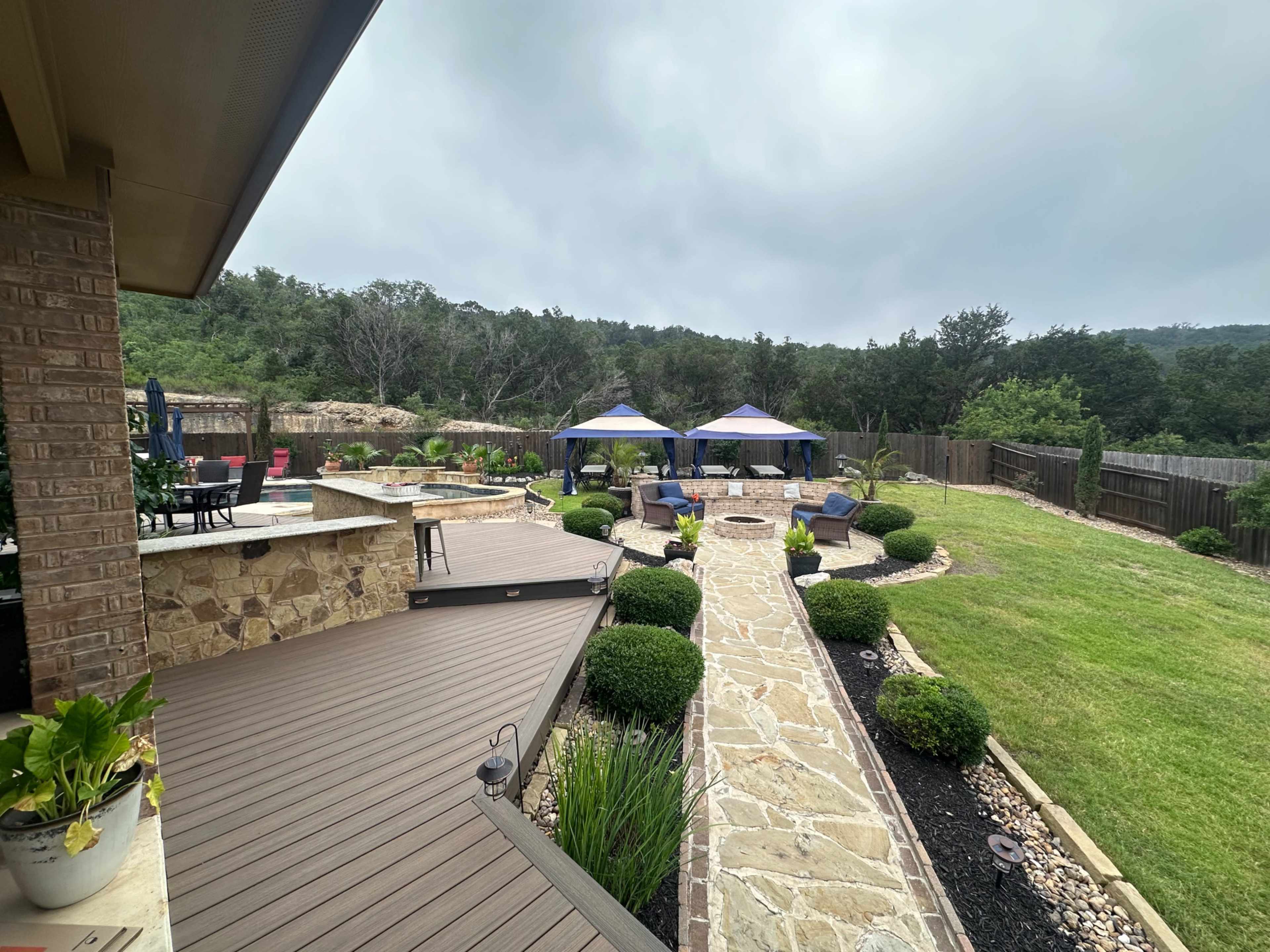 The image shows a landscaped backyard with a stone pathway, several seating areas, and two canopies, set against a backdrop of greenery and cloudy skies.