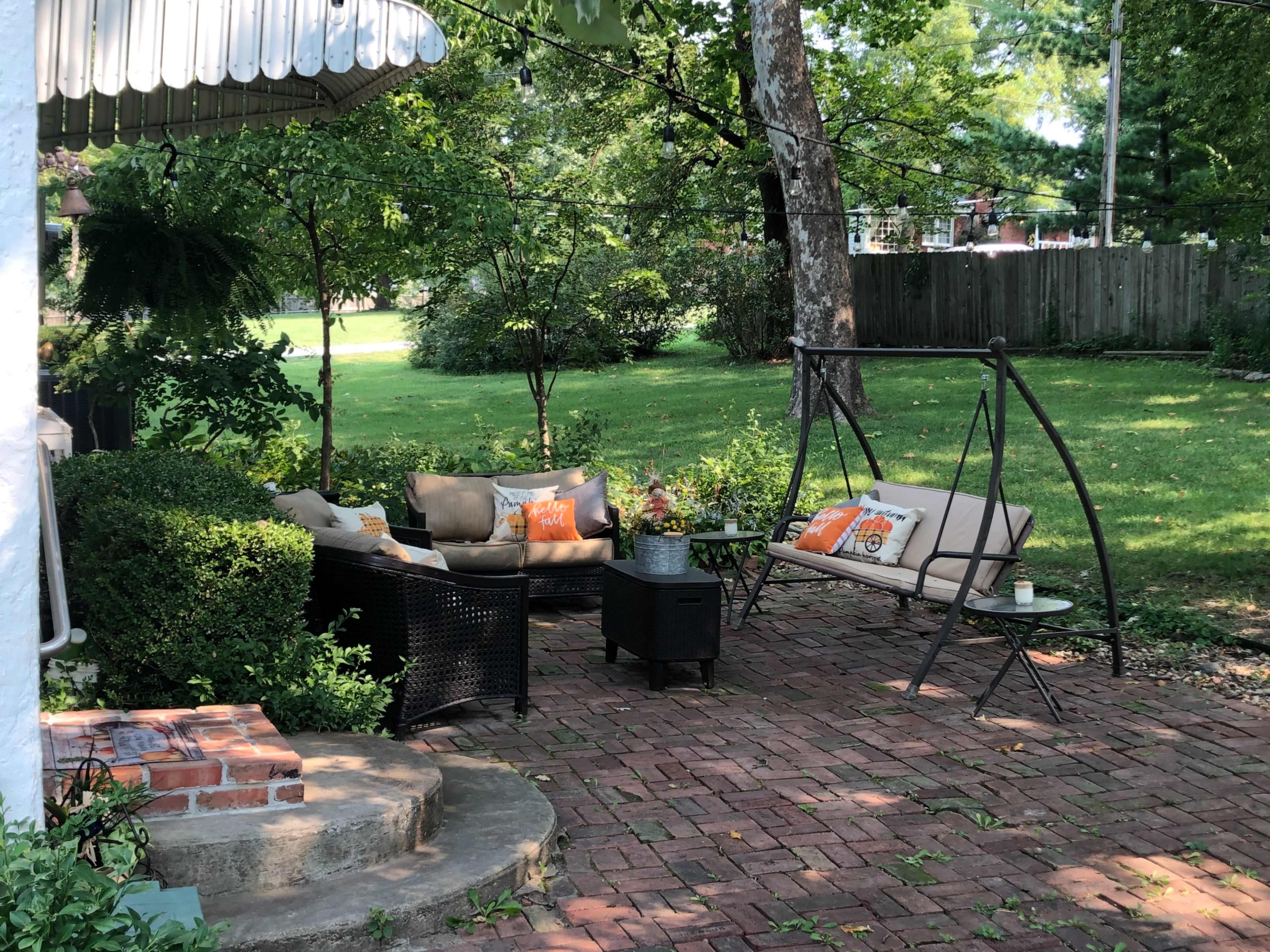 A cozy outdoor seating area features a swing chair and a black wicker sofa surrounded by greenery and brick paving.