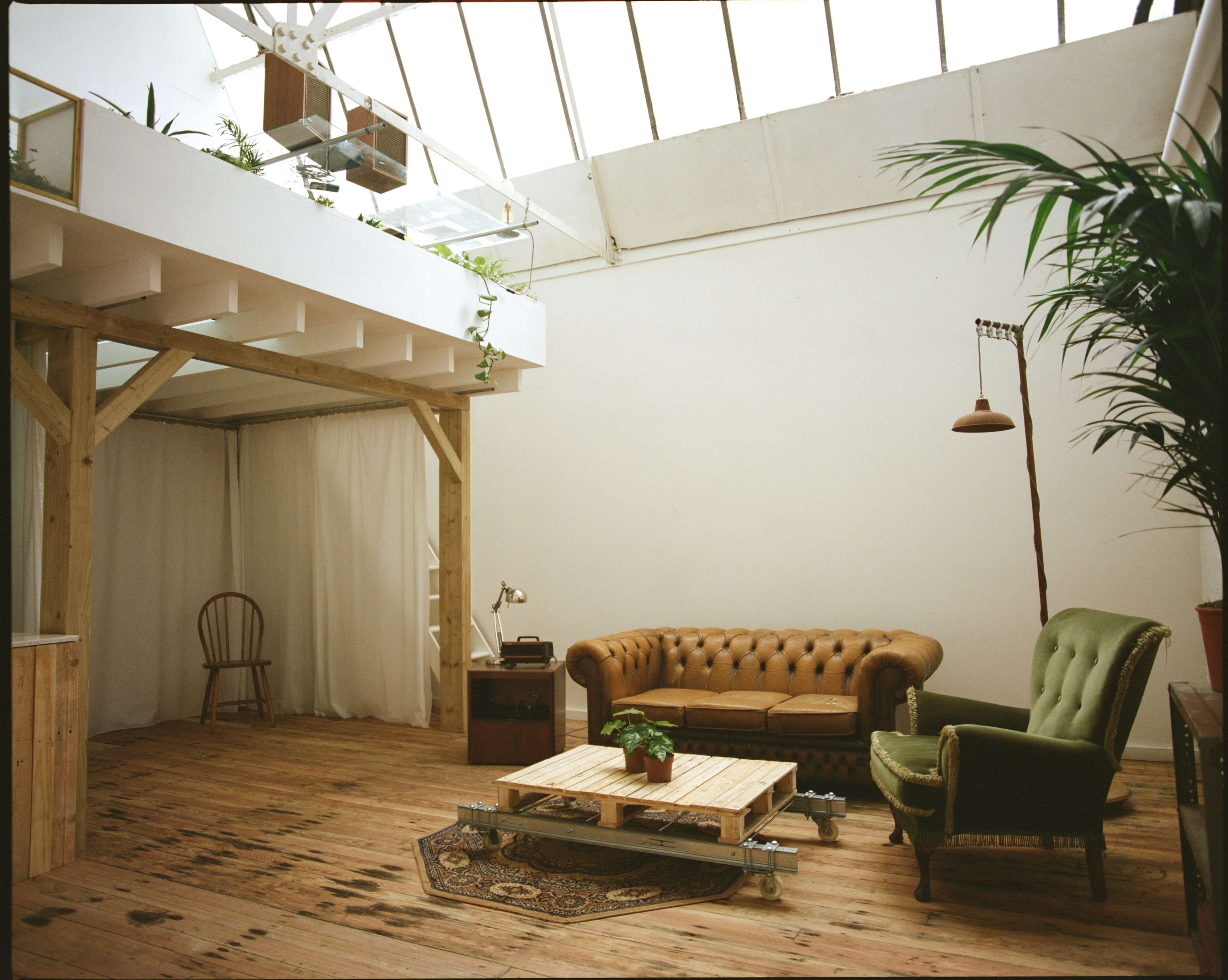 The scene depicts a spacious, well-lit room featuring a wooden floor, a brown leather chesterfield sofa, a green armchair, and a wooden coffee table, with large windows allowing natural light to fill the space.