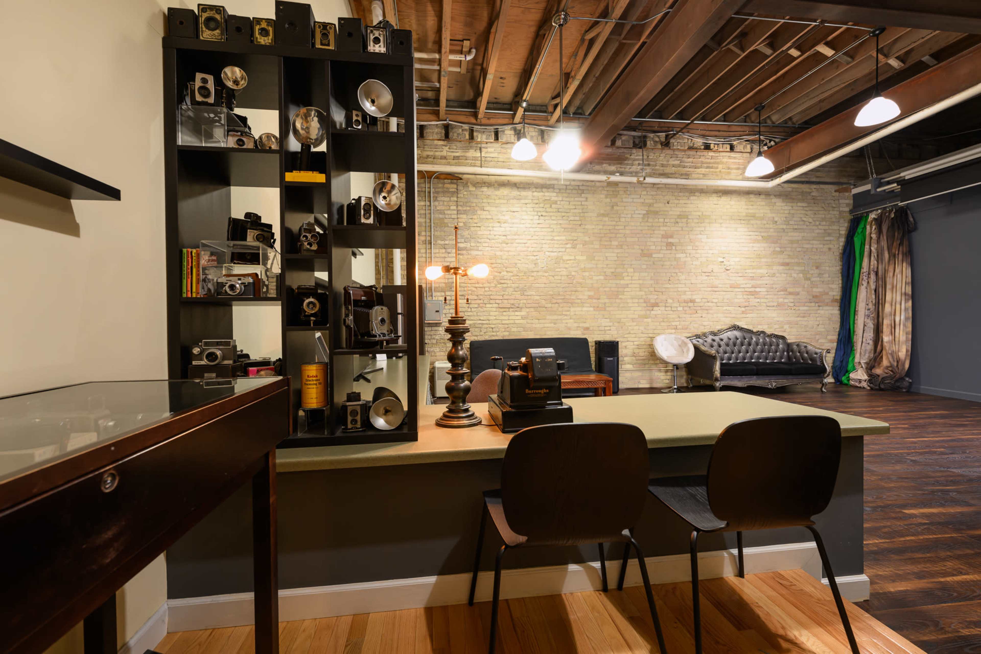 The image shows a well-lit studio space featuring a shelf with vintage cameras and equipment, a desk with two chairs, and a lounge area with a couch and a bed in the background.