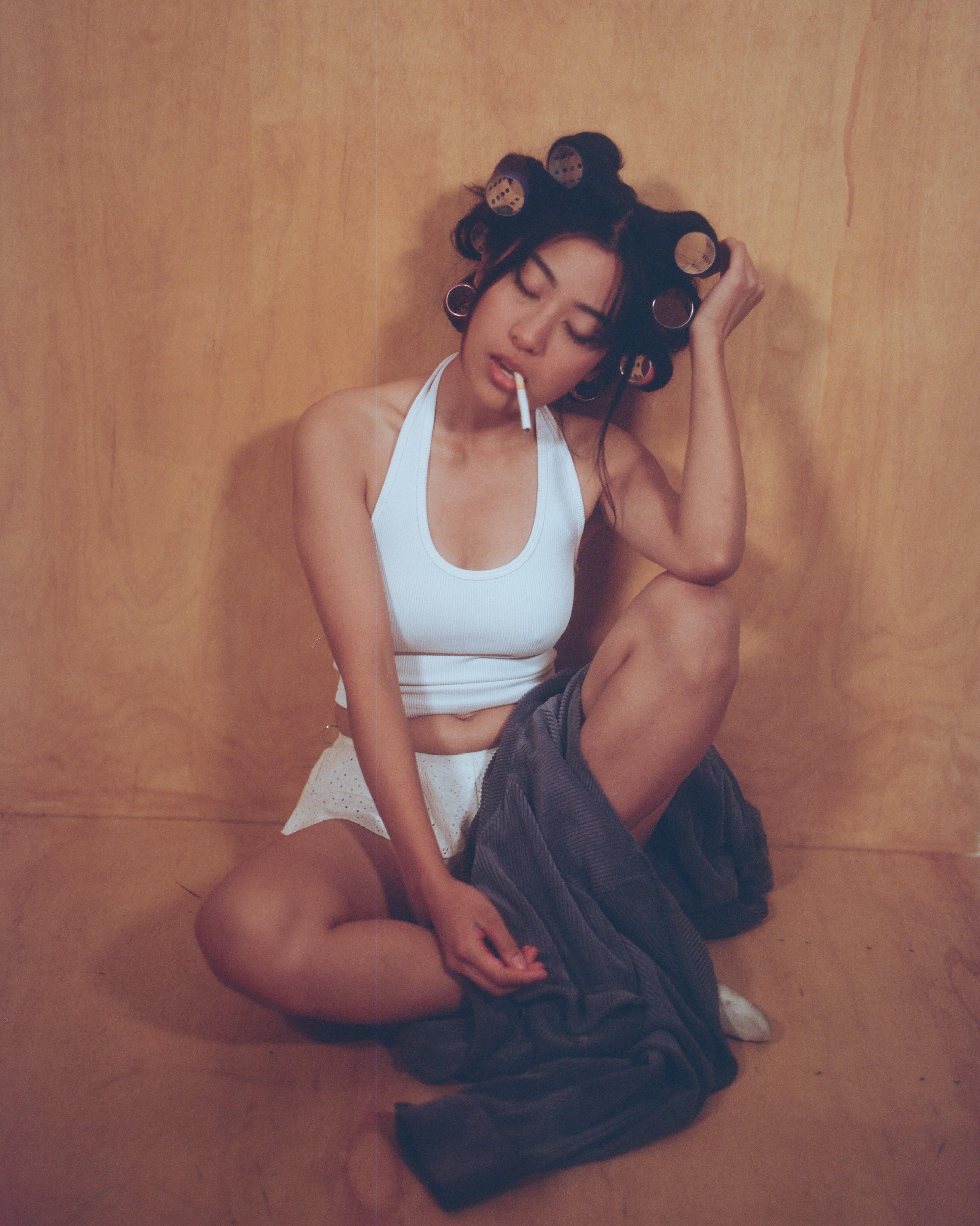 A woman sits on a wooden floor with her legs crossed, wearing a white top and shorts, while holding a cigarette in her mouth and adjusting her hair in rollers.