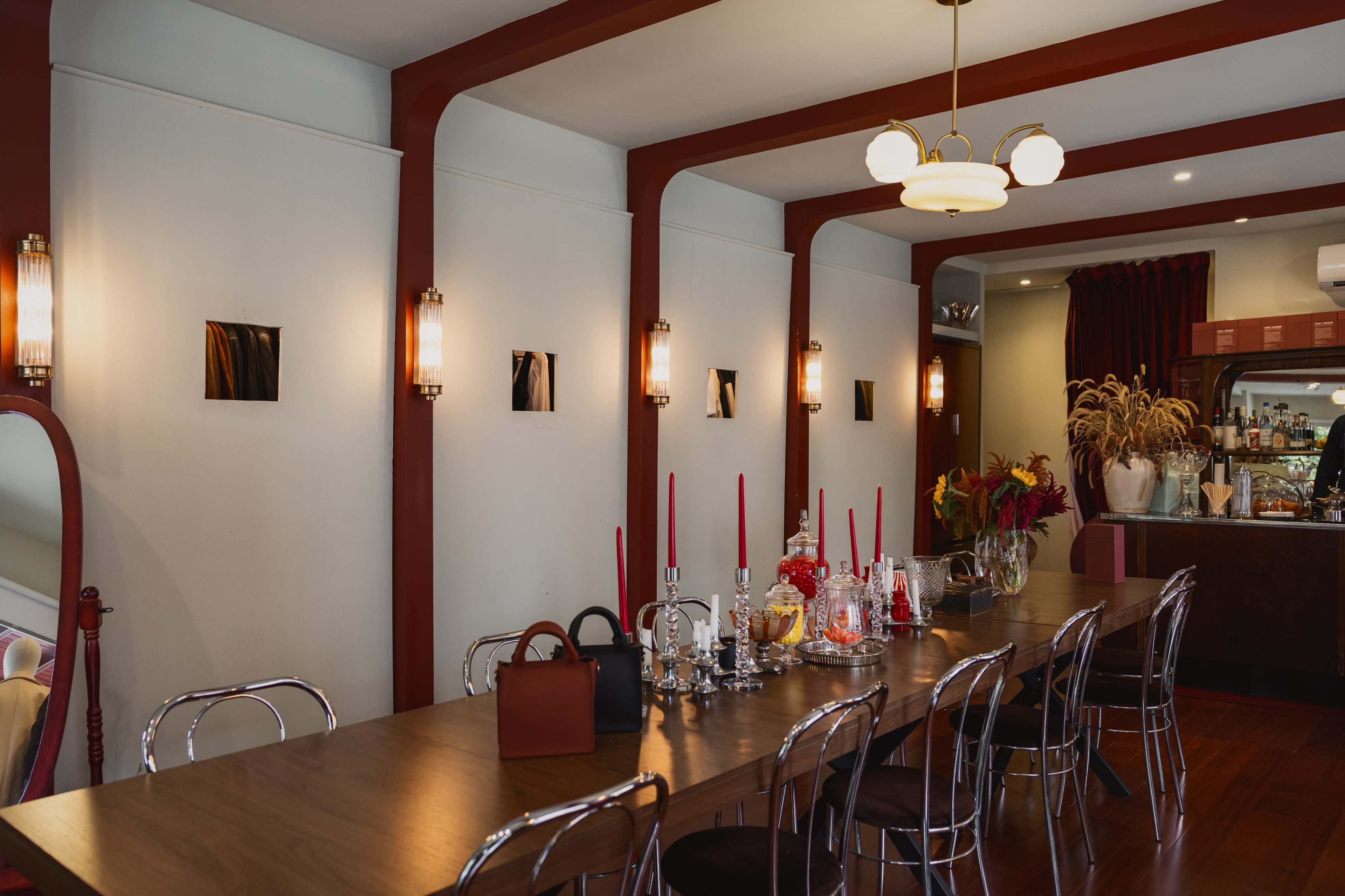 The image shows a dining area with a long wooden table, modern chairs, and decorative elements like candelabras and glassware against a backdrop of red and white walls.