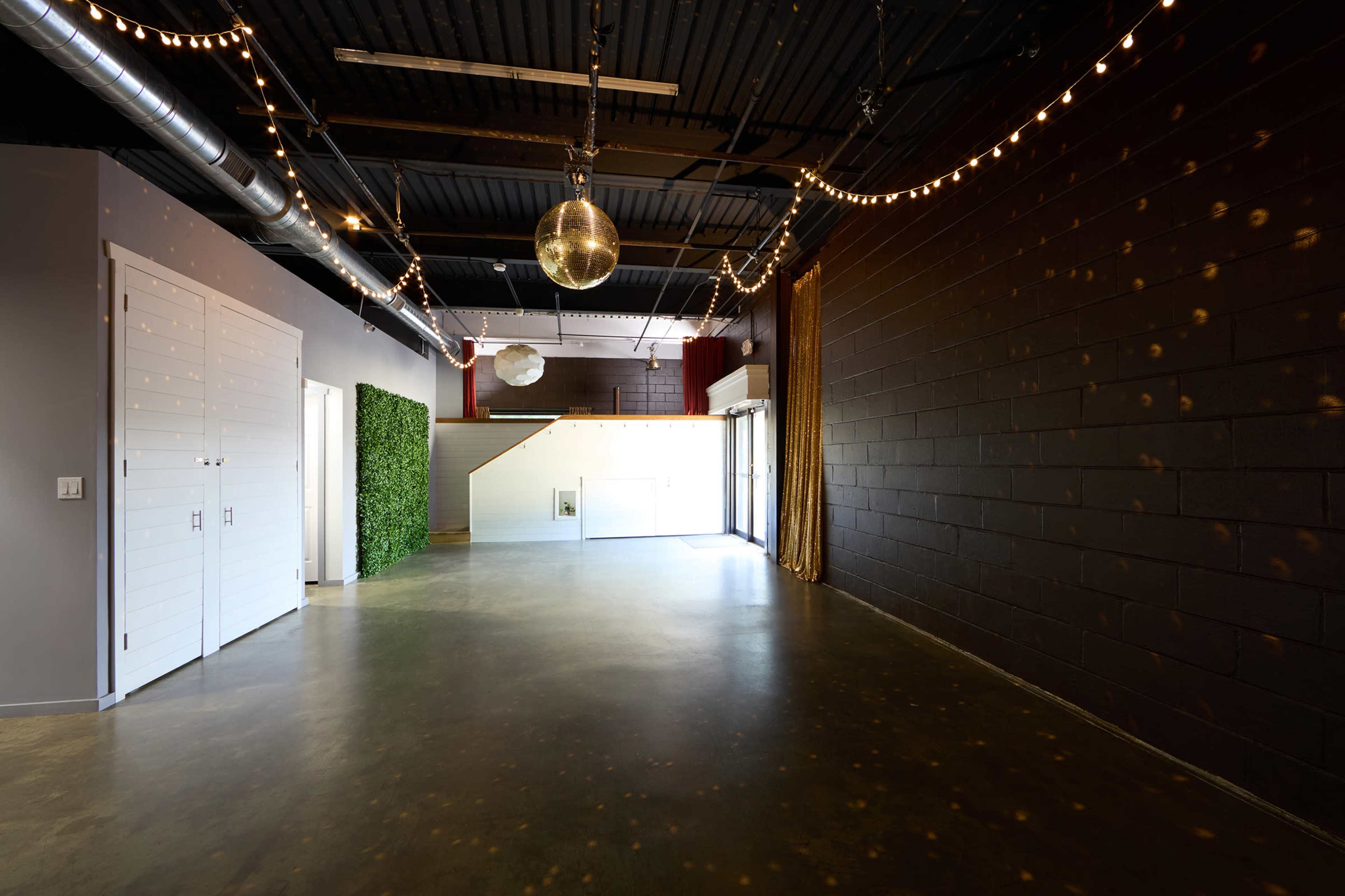 The image shows a spacious interior venue with a polished concrete floor, adorned with string lights and a disco ball hanging from the ceiling, alongside a wall covered in dark paint and a section with greenery.
