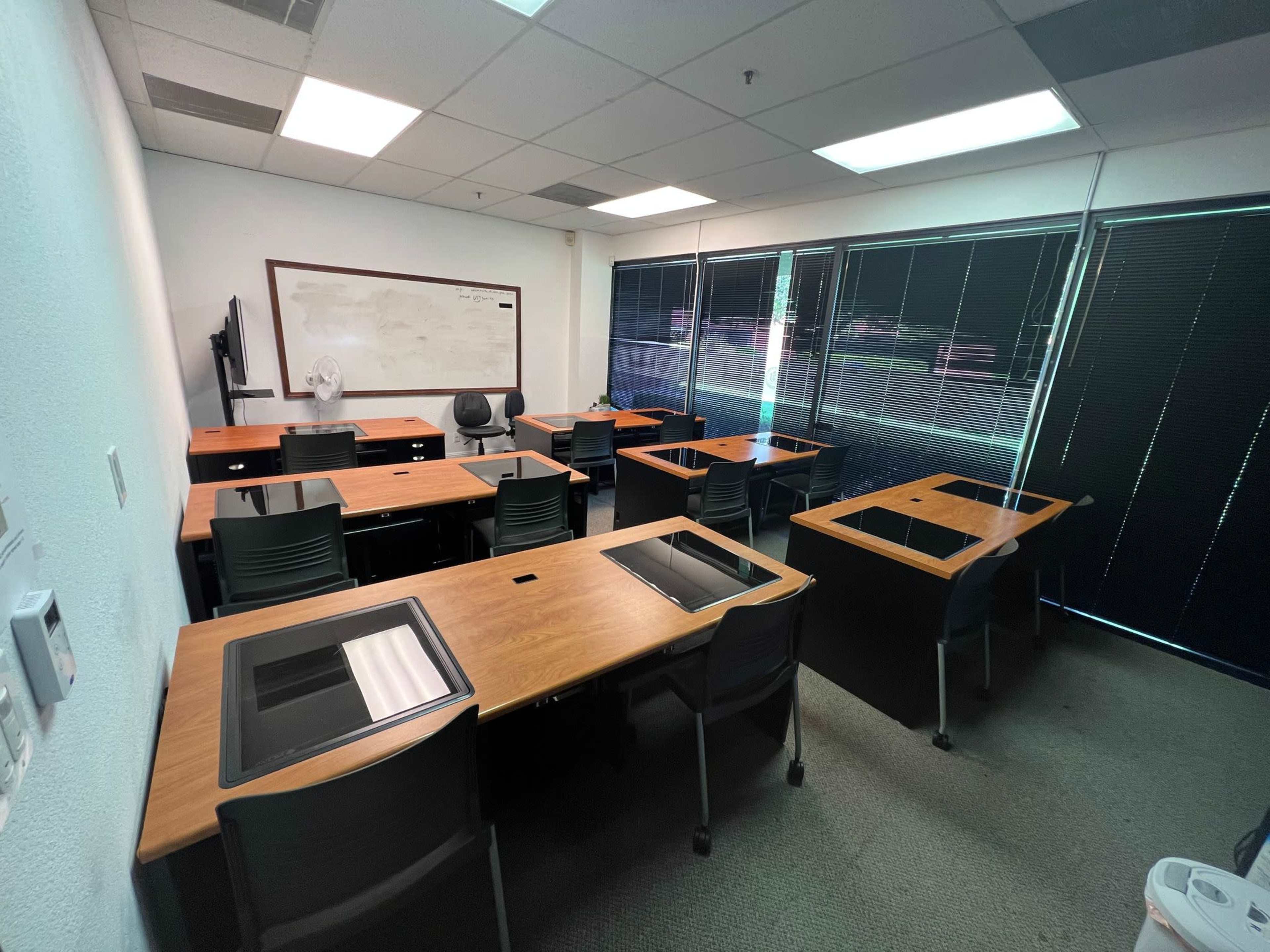 The image shows a sparsely populated classroom with wooden desks arranged in a grid and large windows covered by blinds.