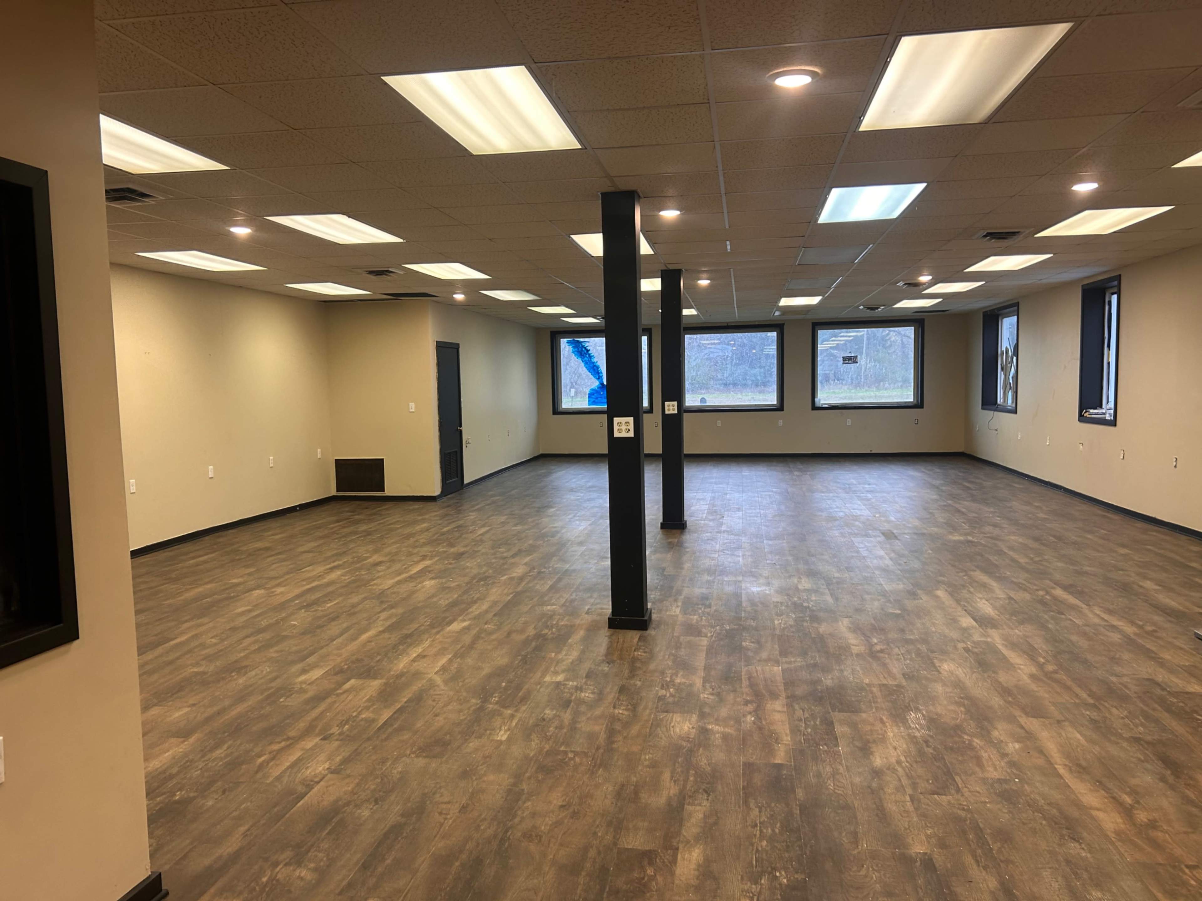 An empty room with light-colored walls and a wood-patterned floor features several windows and black pillars supporting the ceiling.