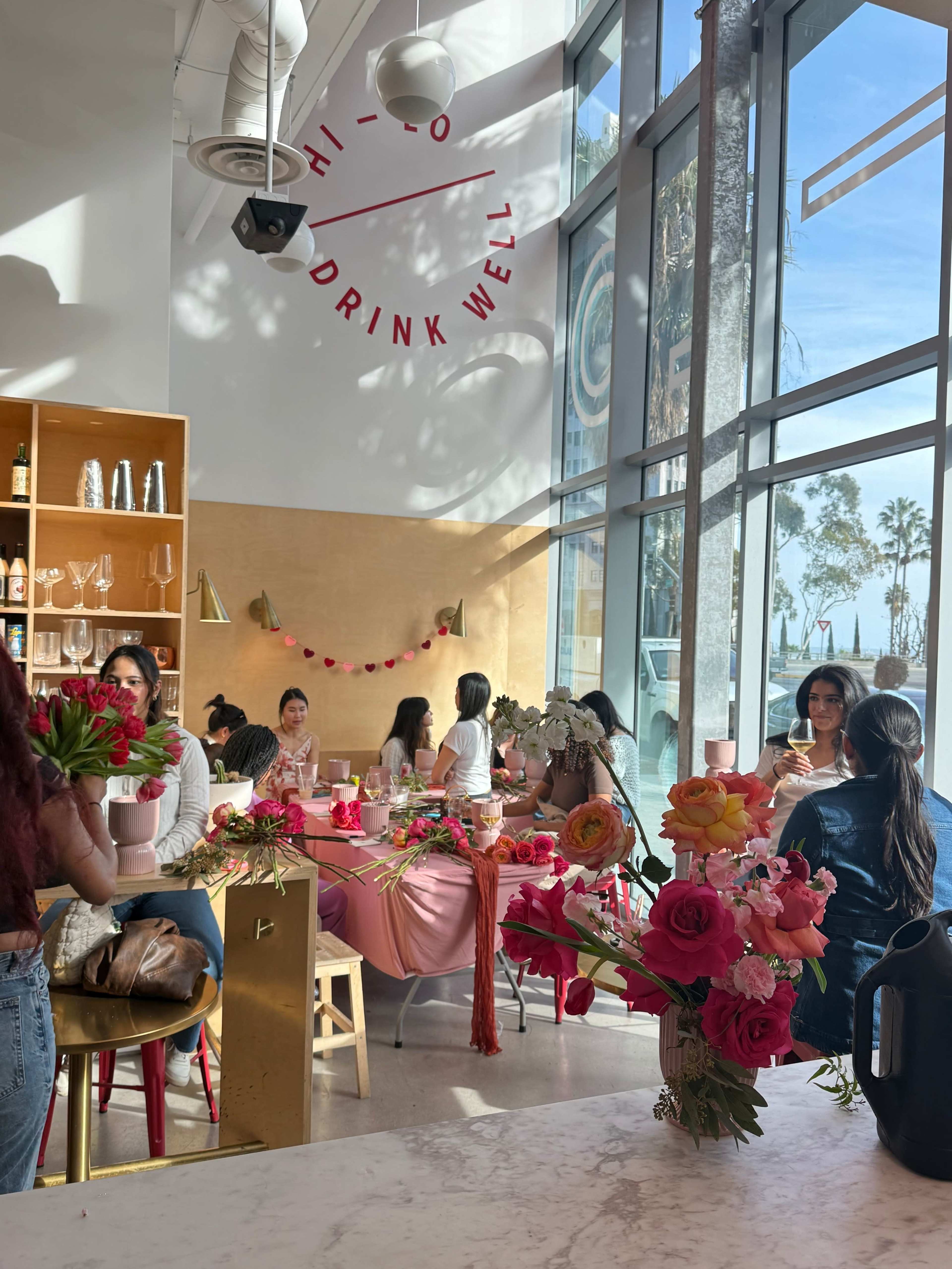 A bright, modern cafe features several tables decorated with flowers and pink tablecloths, while customers enjoy drinks and socialize near large windows displaying palm trees outside.