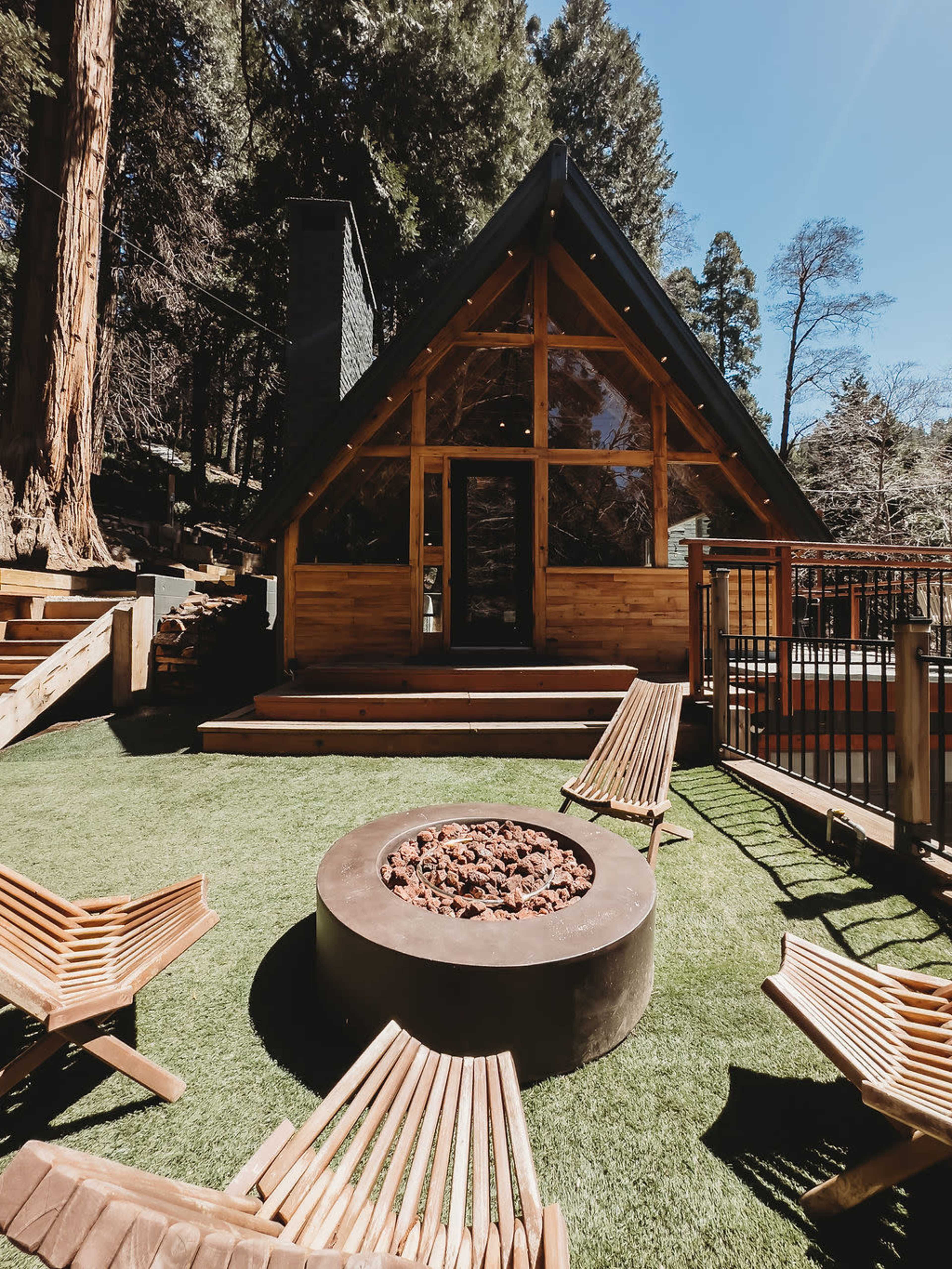 A wooden cabin with a triangular roof is surrounded by seating and a fire pit on a grassy area.