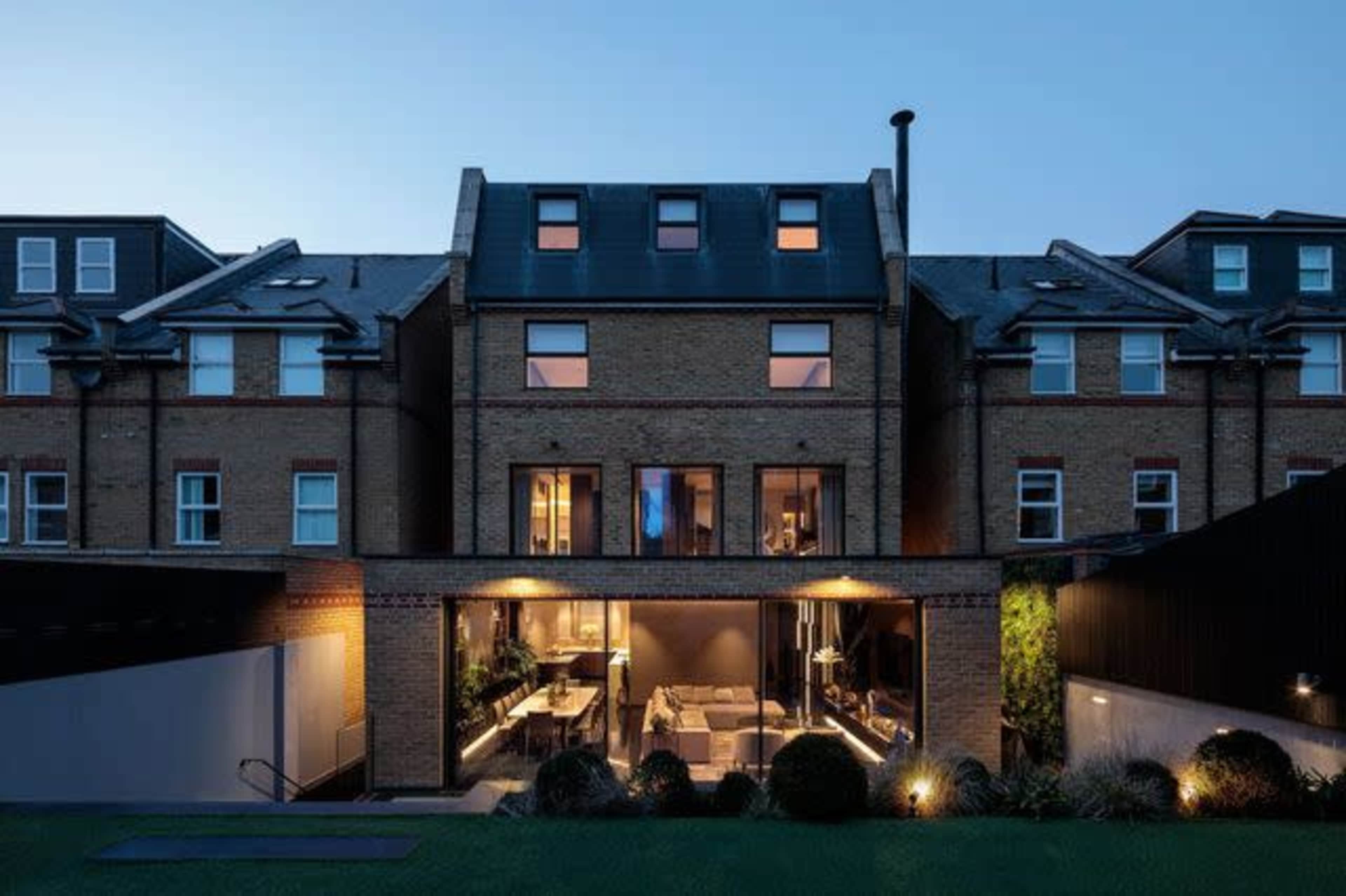 The image shows a modern two-story brick house with large windows and a patio visible in the evening light, framed by other similar buildings.