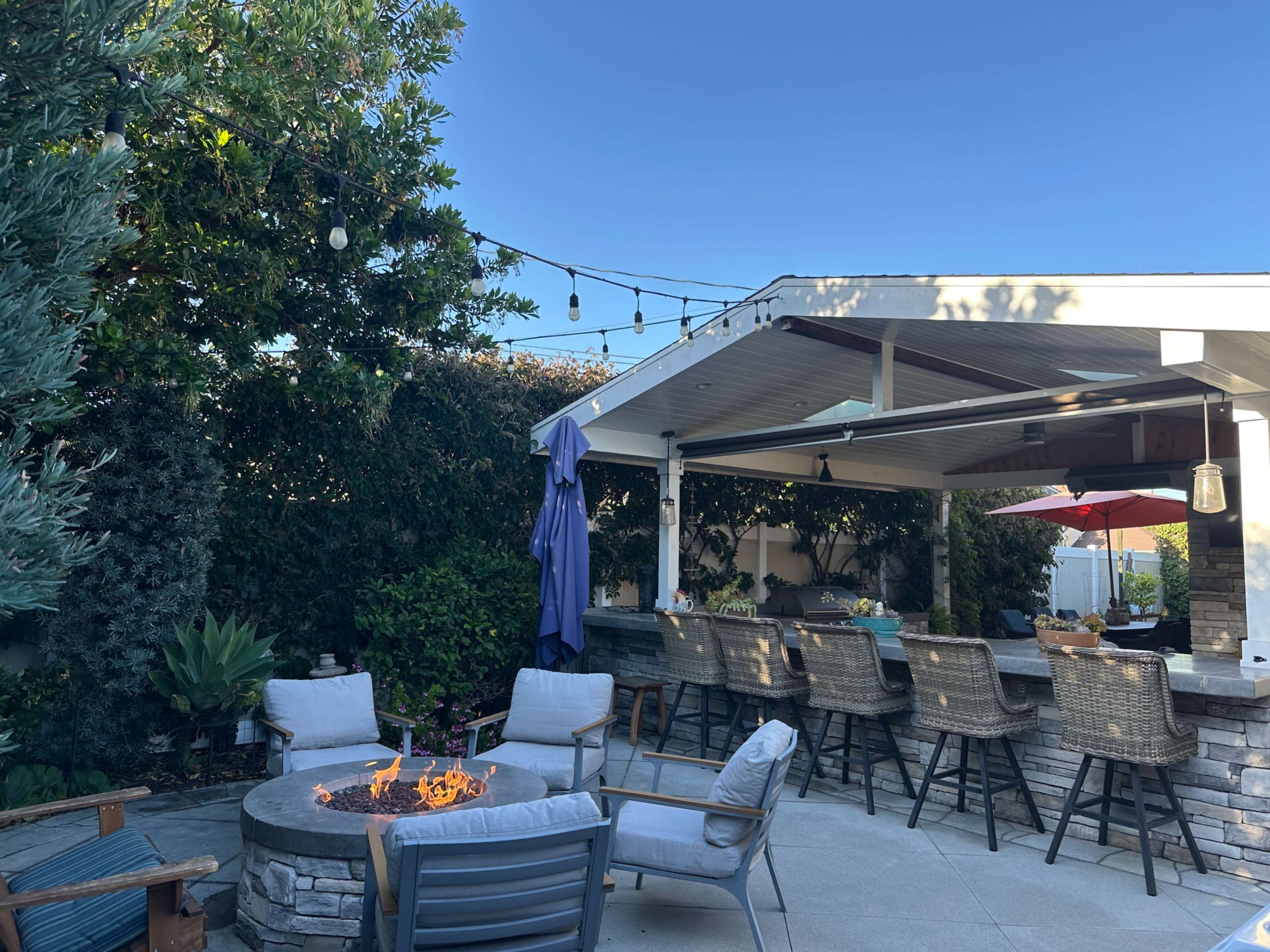 The image shows a patio area featuring a circular fire pit surrounded by seating, with a bar and outdoor kitchen under a covered structure, and string lights hanging above.