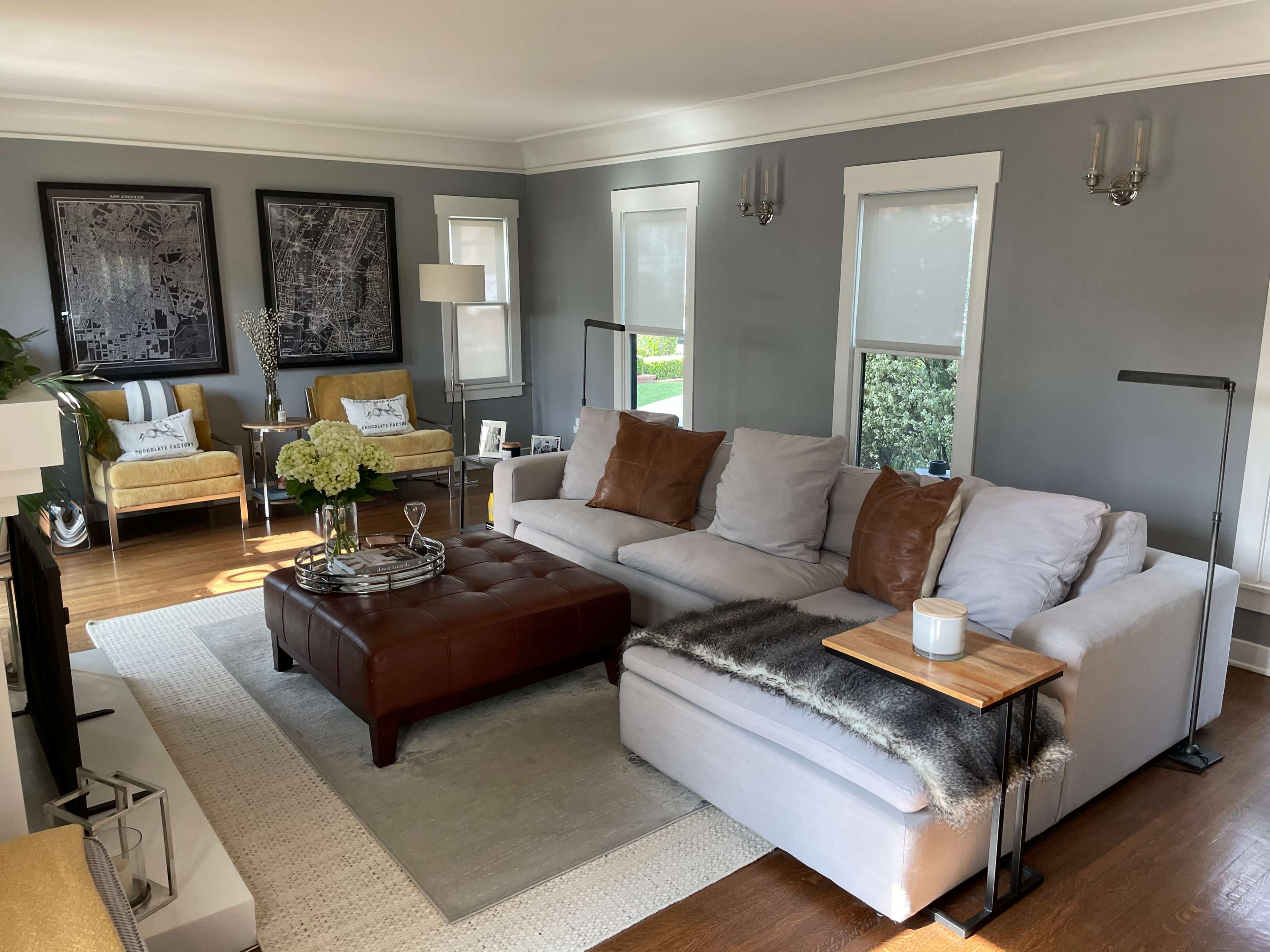 The living room features a light gray sectional sofa, a brown leather ottoman, and two yellow armchairs, with framed black and white maps on the walls.