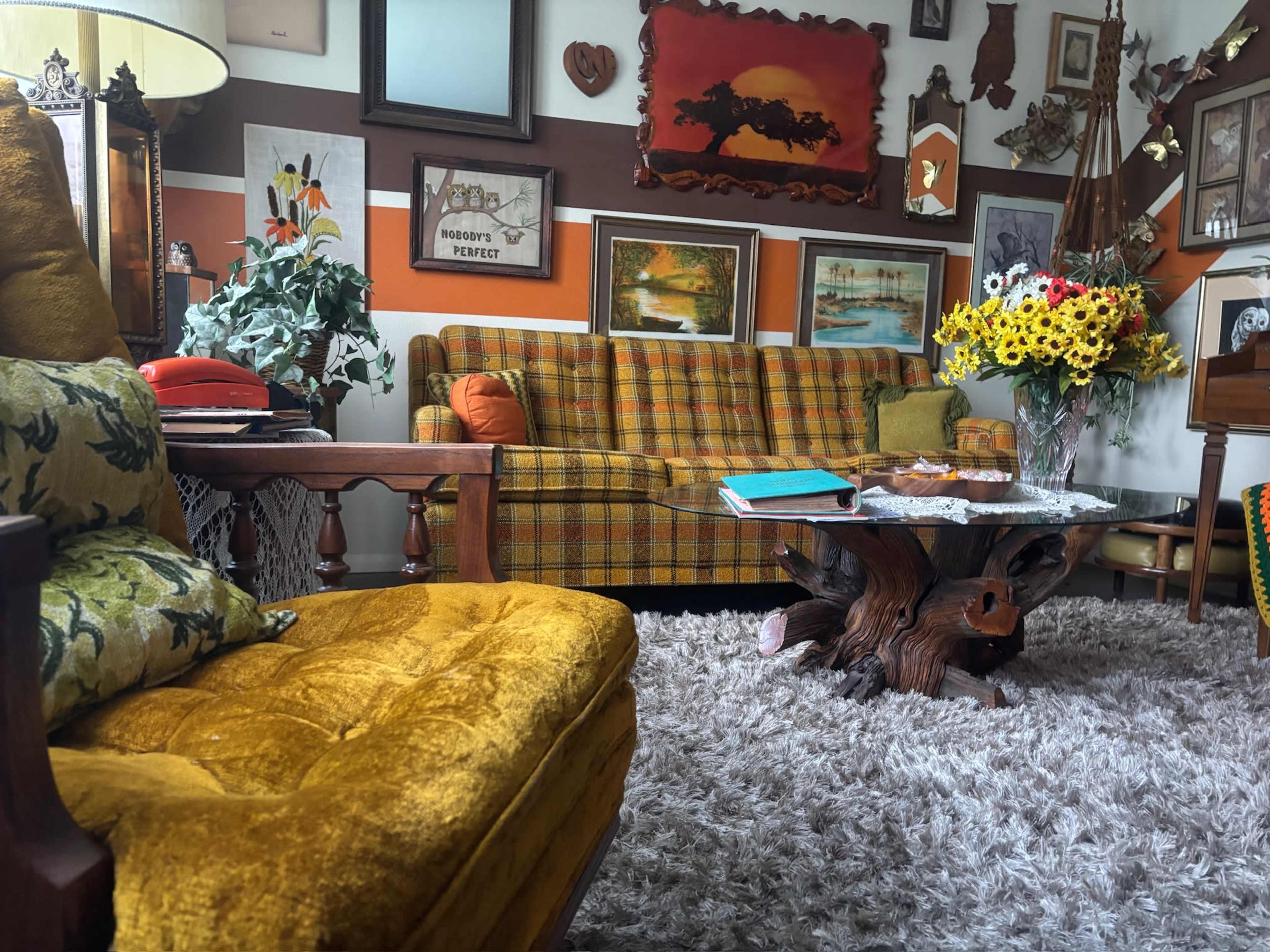 The image shows a vintage living room with a checkered sofa, a wooden coffee table, and various framed artworks and decorations on the walls.