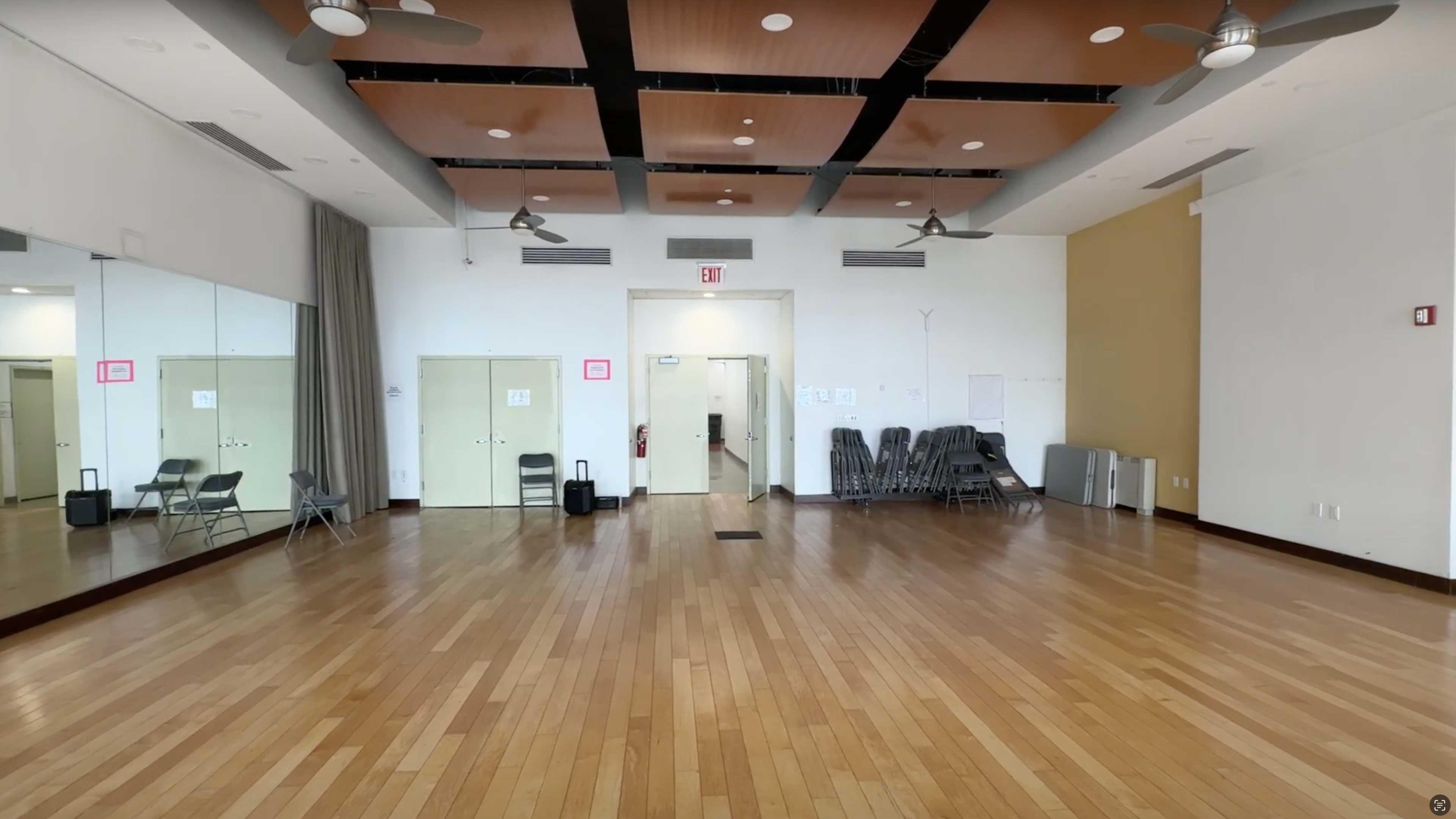 An empty room with wooden flooring, ceiling fans, and a mirrored wall, featuring chairs stacked against the side and double doors at the far end.