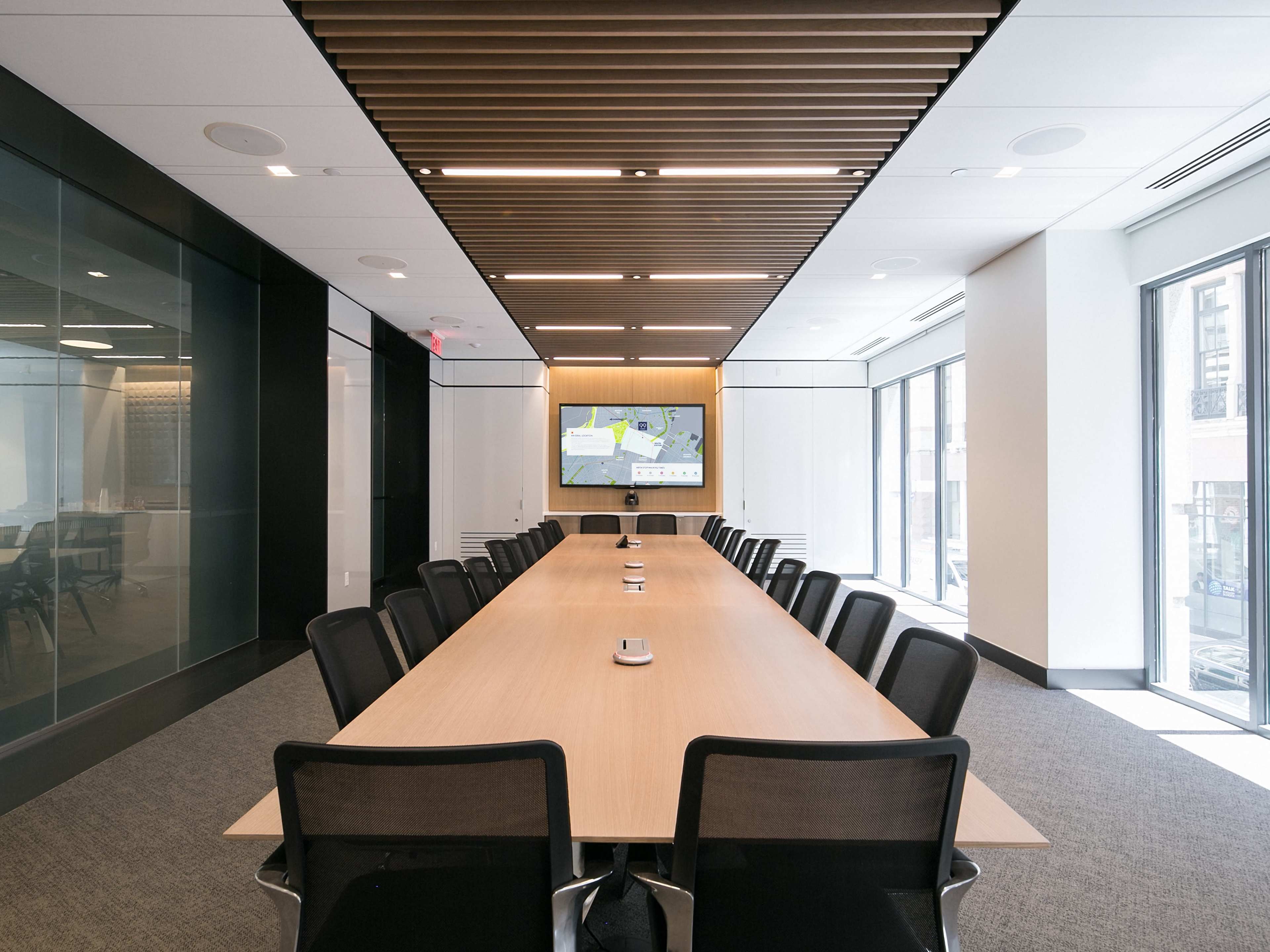 A modern conference room features a long wooden table surrounded by black mesh chairs, with large windows and a screen displaying a presentation on one wall.