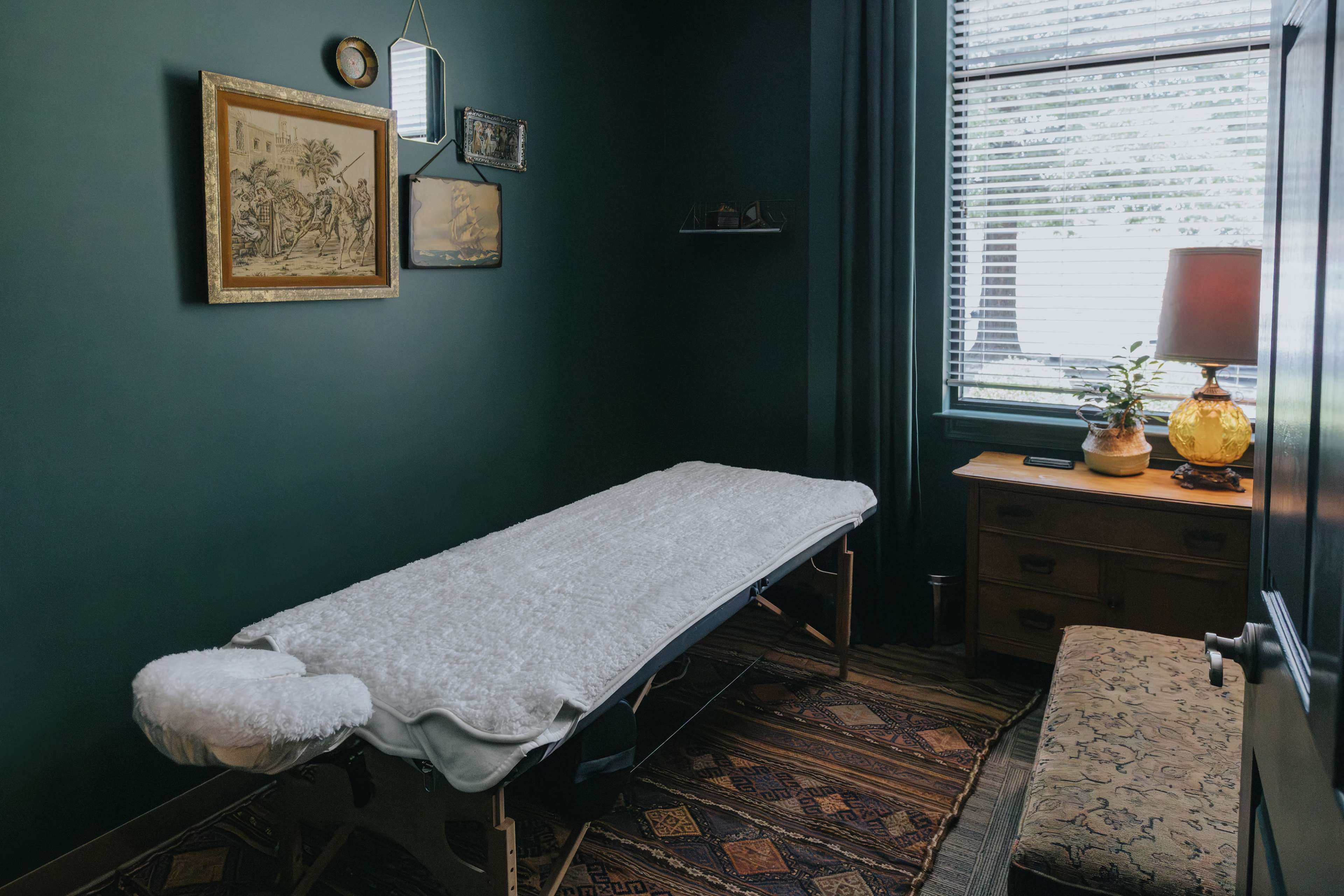 A massage room with a massage table covered in a white sheet, a side table with a lamp, and framed artwork on the walls.