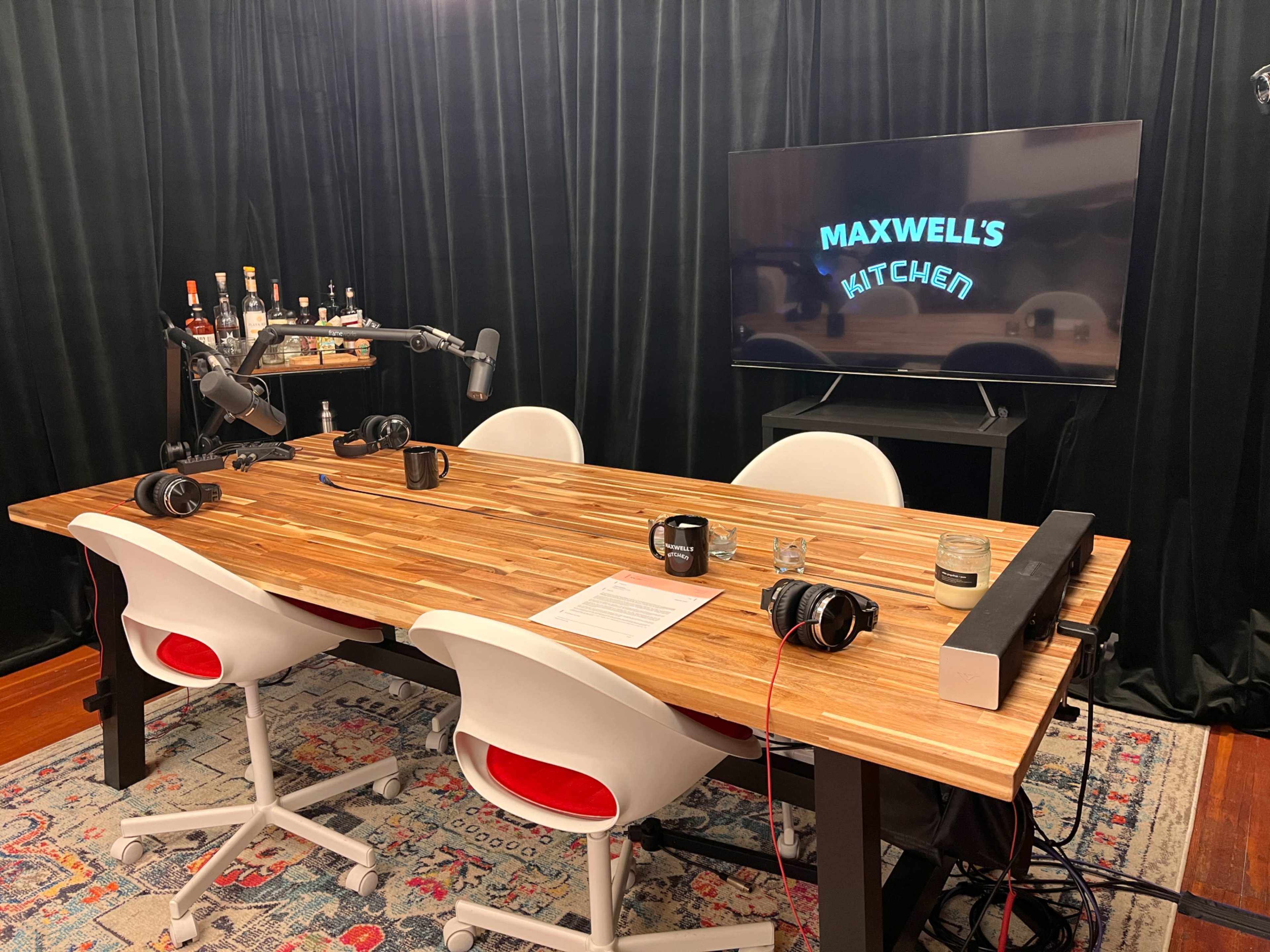 The image shows a modern podcast studio featuring a wooden table, white chairs, microphones, and a television displaying "MAXWELL'S KITCHEN," with a black curtain in the background.