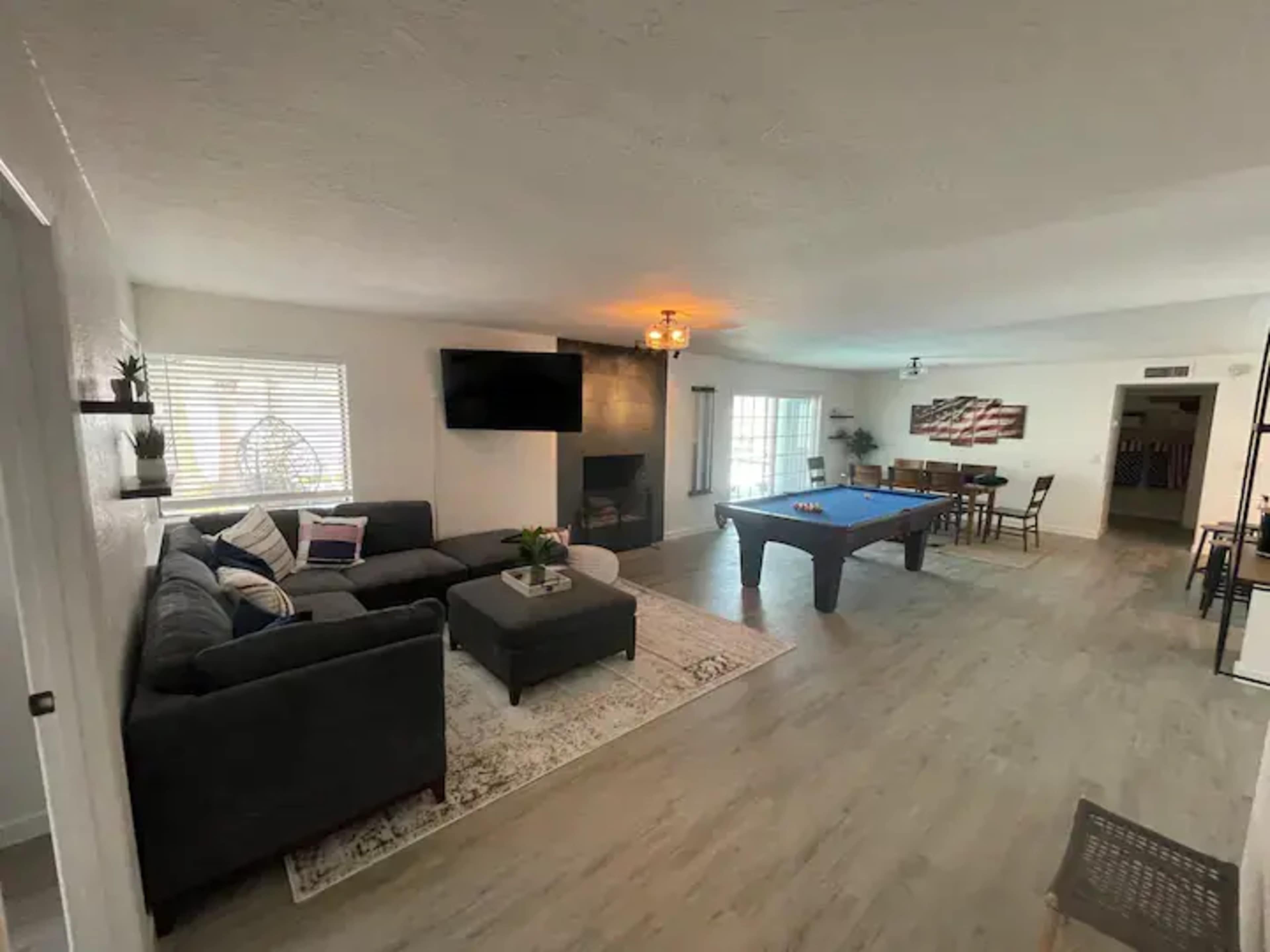 A spacious living room with a grey sectional sofa, a pool table, a dining area, and a large window providing natural light.