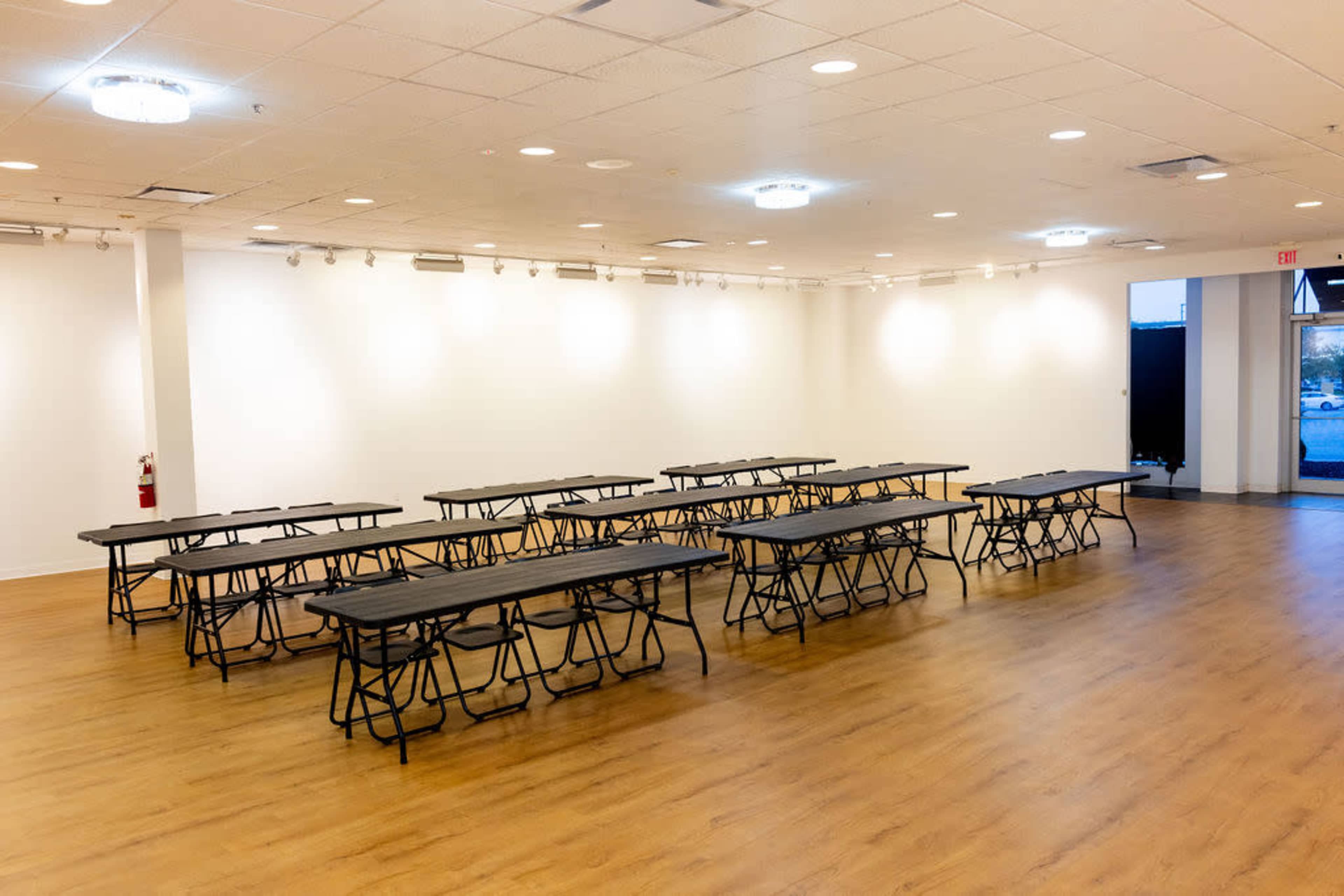 The image shows a spacious room with multiple black folding tables arranged in rows on a wooden floor, illuminated by overhead lights.