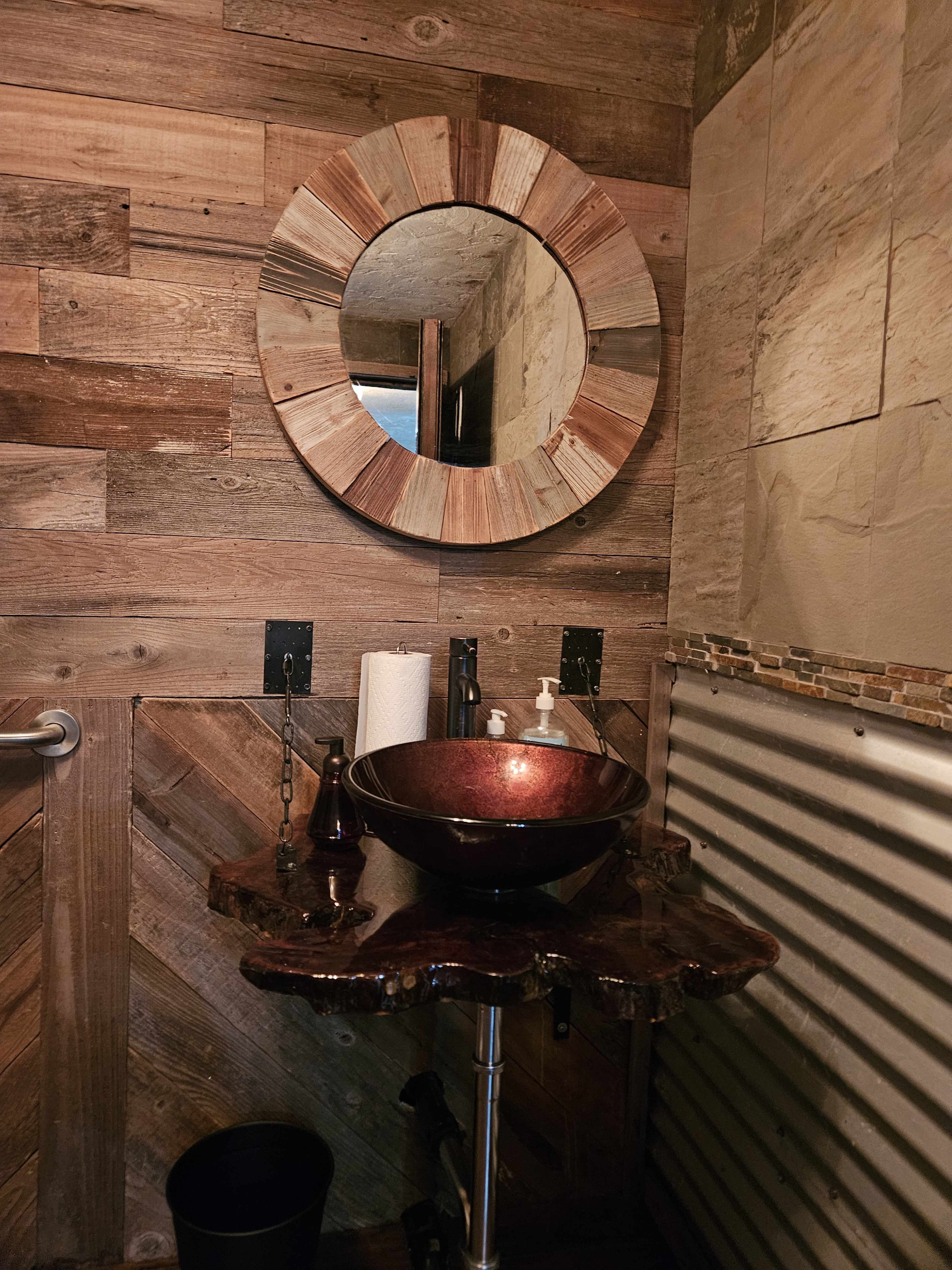 The image shows a rustic bathroom setting featuring a round mirror, a dark bowl sink on a wooden countertop, and wood-paneled walls.