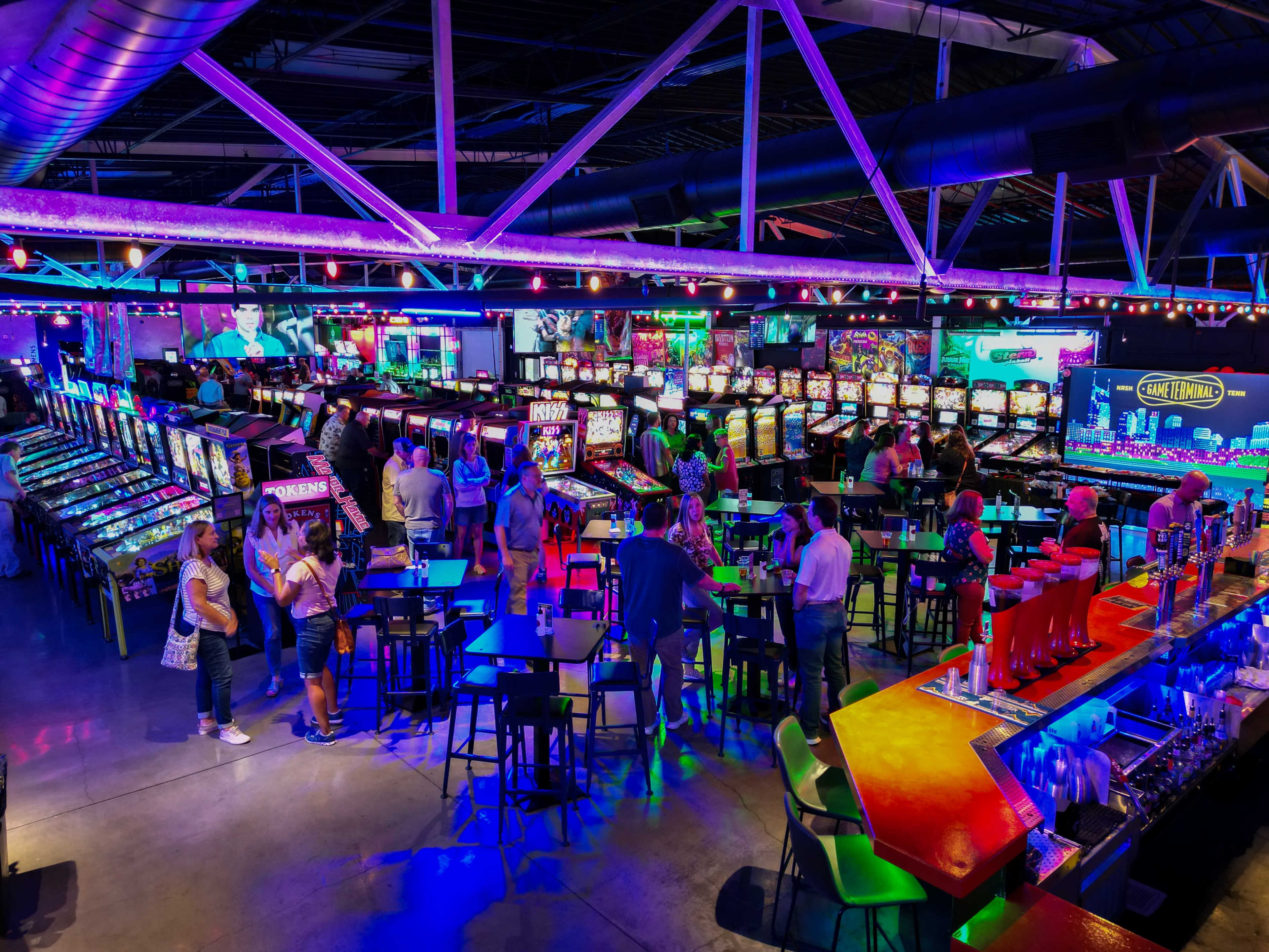 The image shows a lively arcade with rows of colorful slot machines, patrons socializing at tables, and a bar area illuminated by vibrant lights.