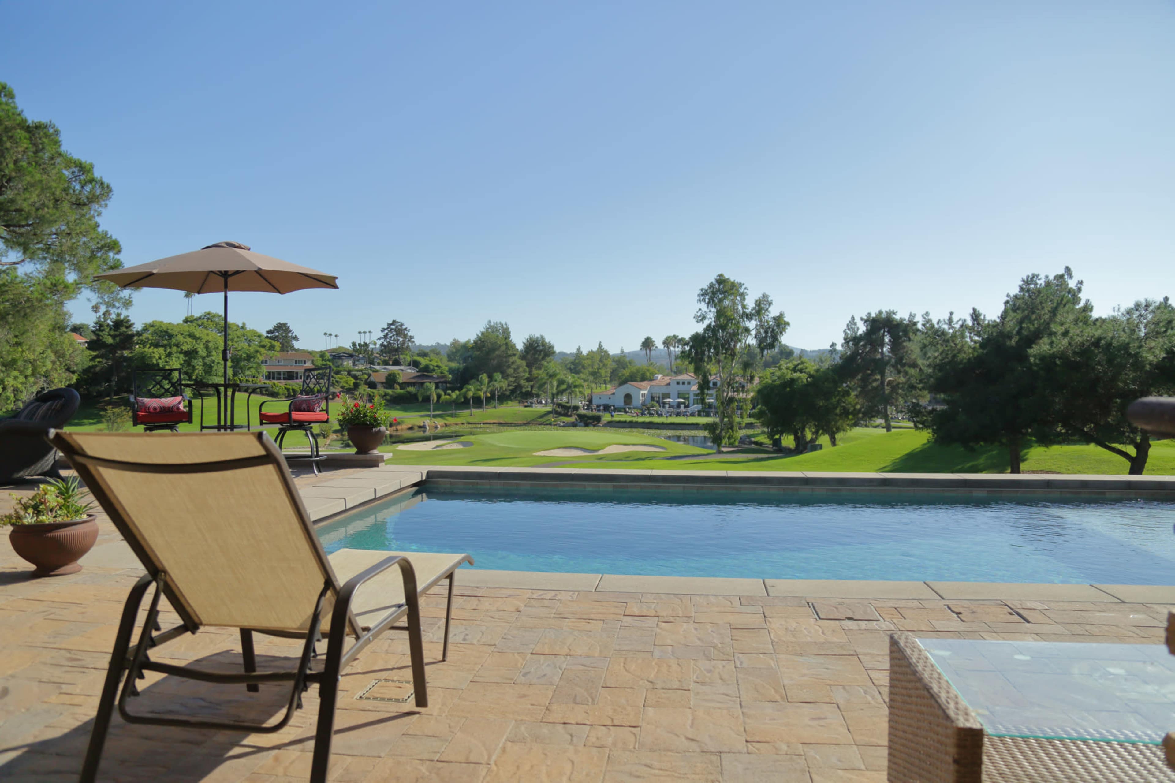 A lounge chair is positioned beside a swimming pool that overlooks a green golf course with trees and a clubhouse in the background.