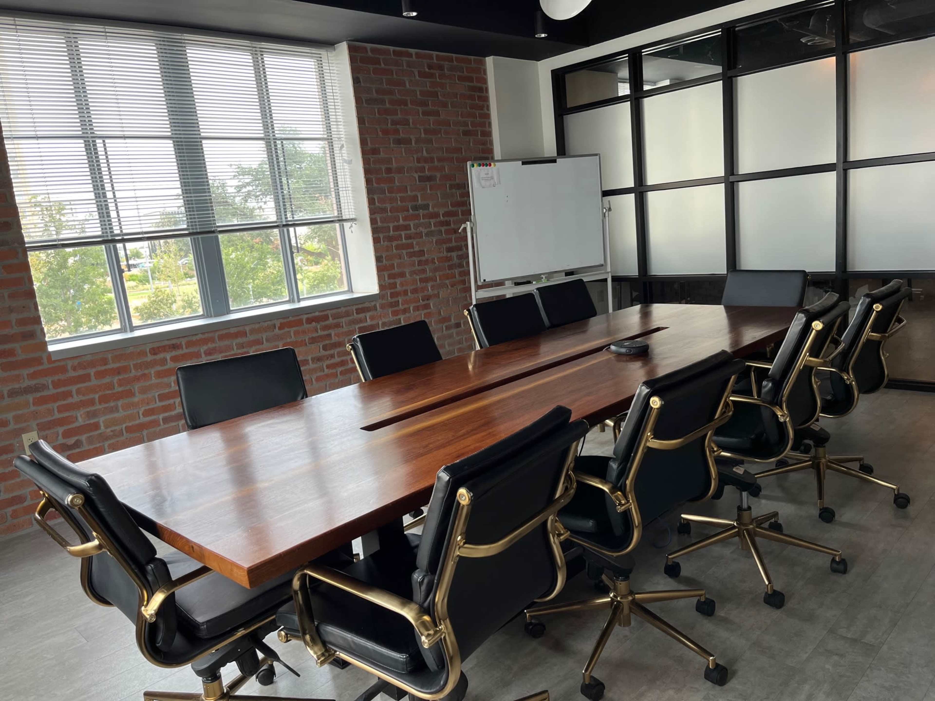 A large wooden conference table is surrounded by black office chairs in a bright meeting room with brick walls and large windows.