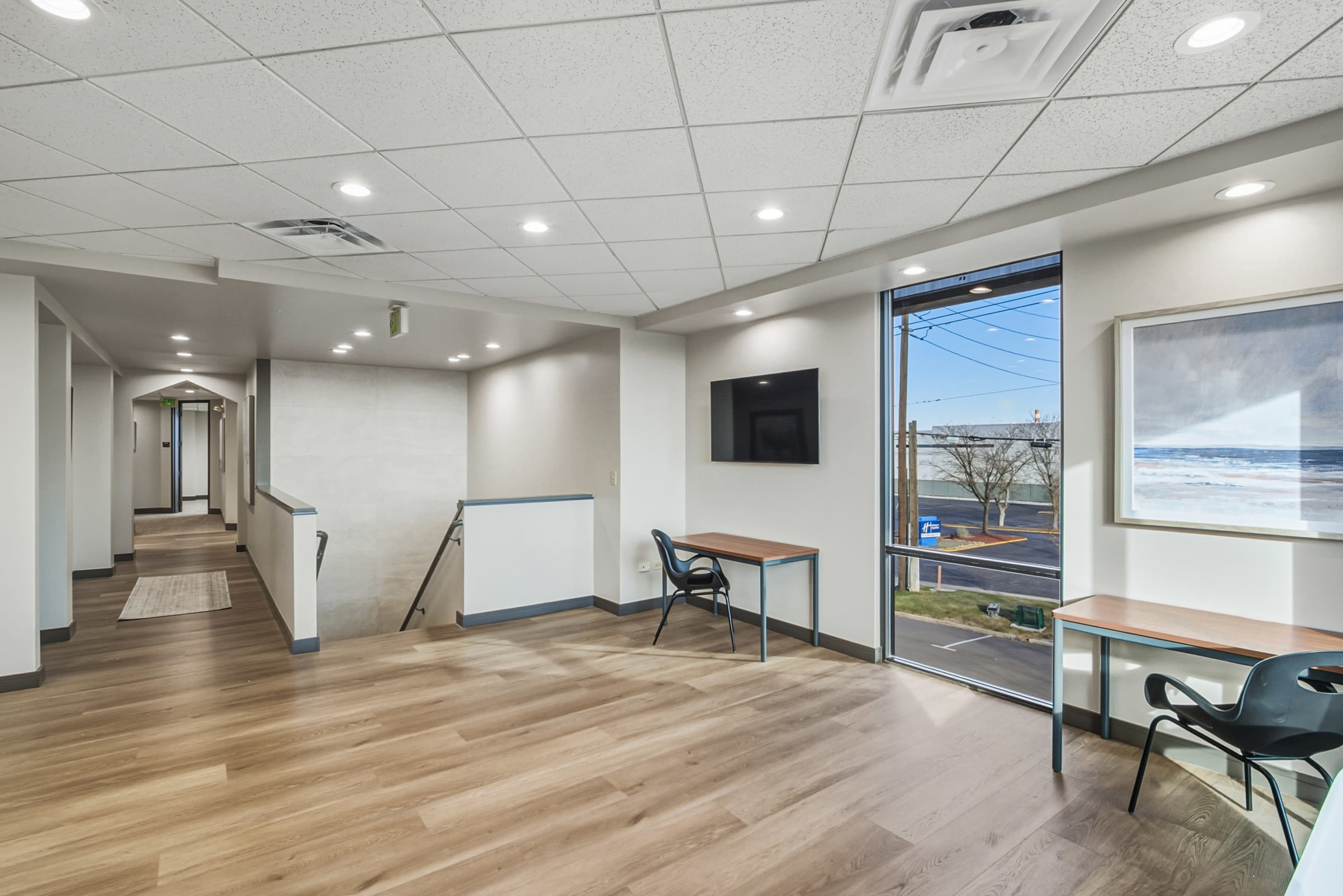 The image shows a spacious interior office area featuring two tables and chairs, a television mounted on the wall, and large windows allowing natural light to enter.