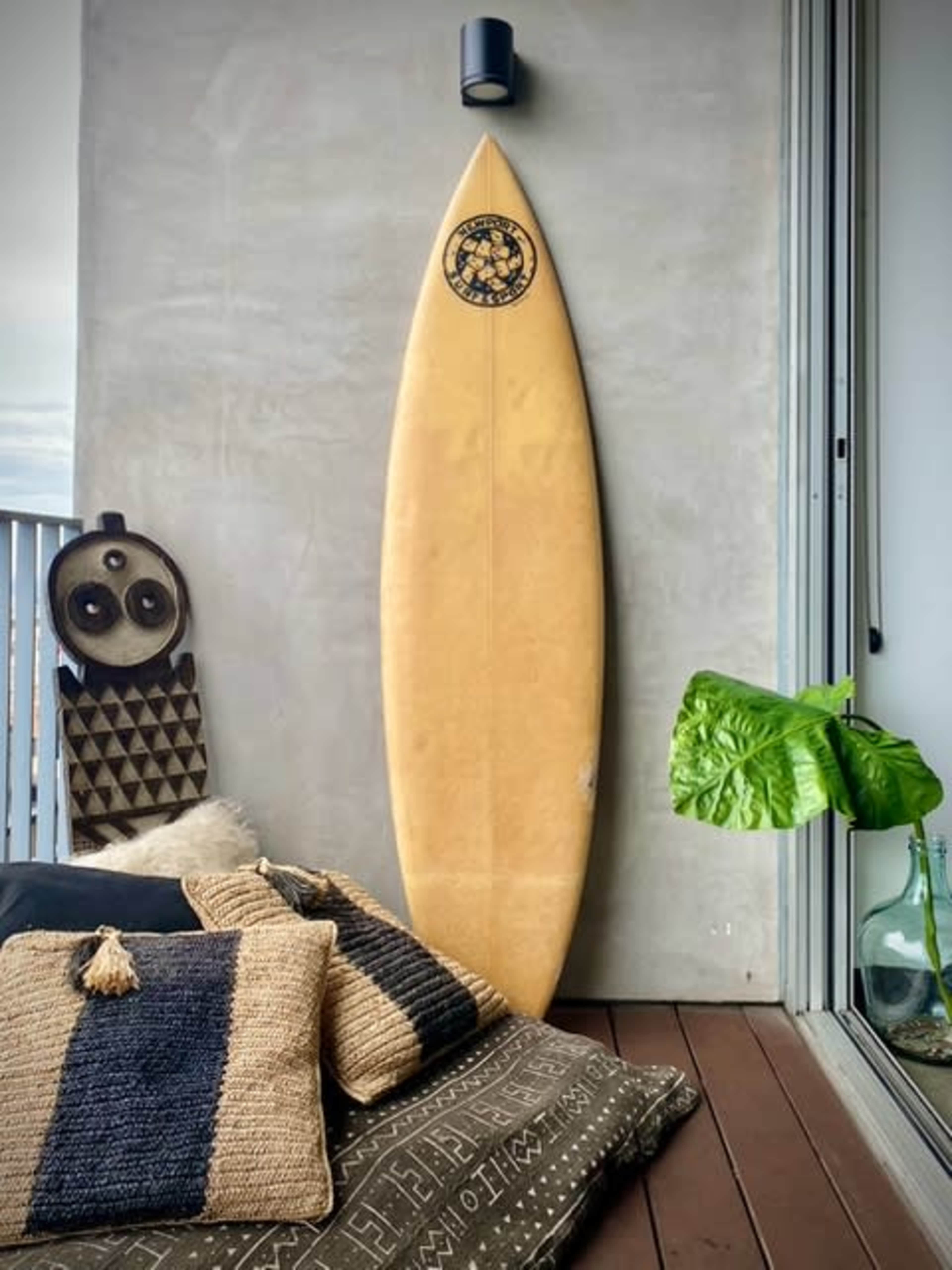 A vintage surfboard leans against a concrete wall beside textured cushions and a large green plant on a wooden deck.
