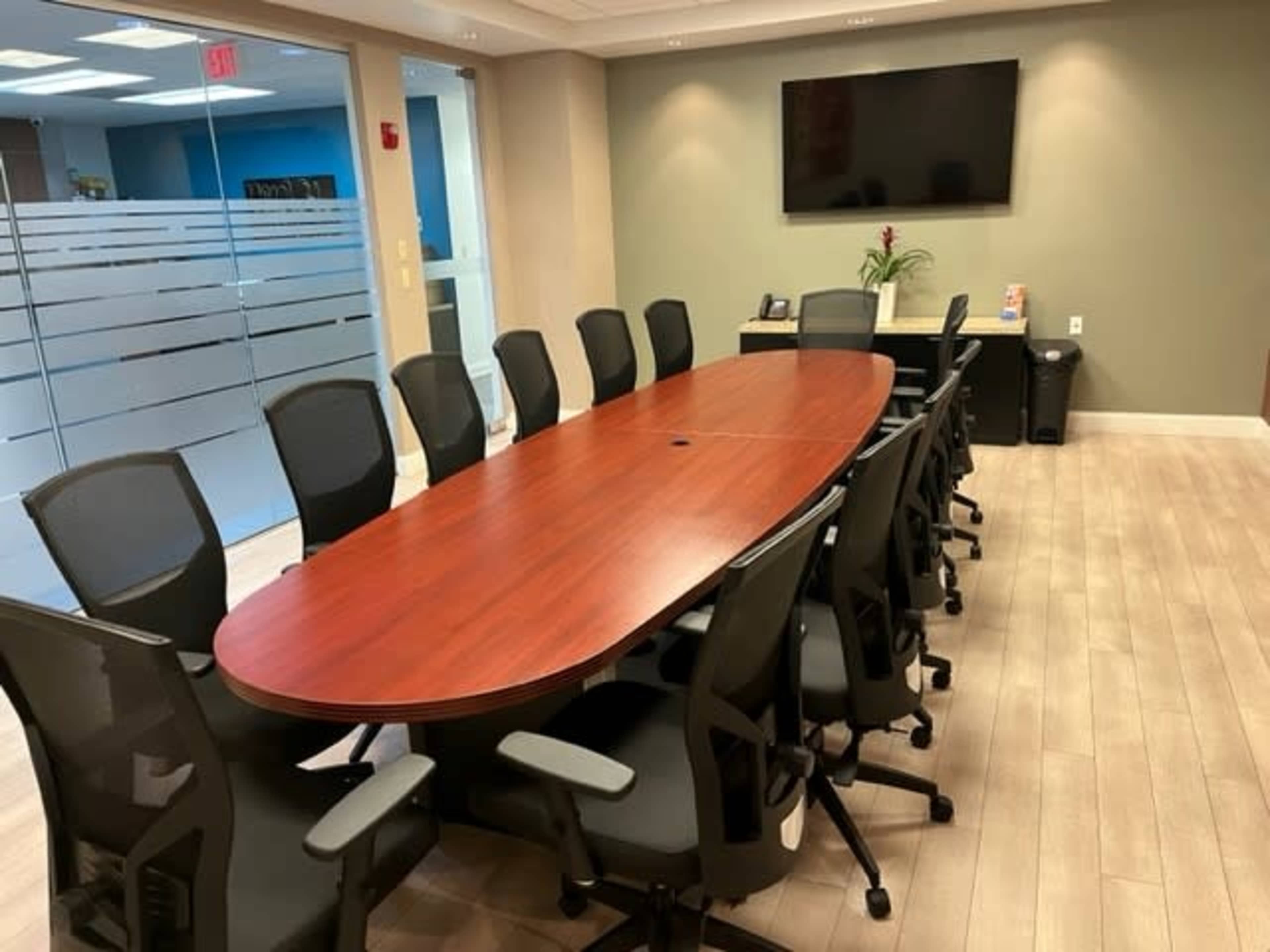 A large, oval conference table is surrounded by black ergonomic chairs in a modern meeting room with a television mounted on the wall.