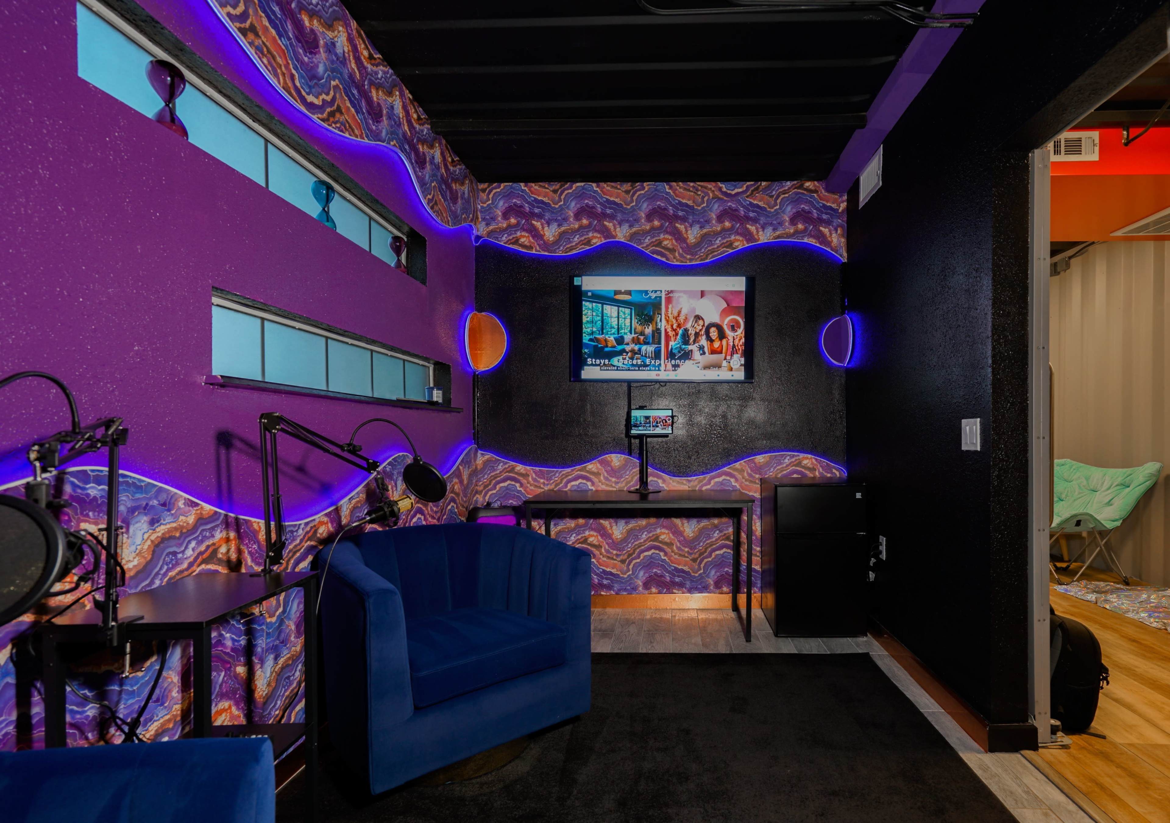 The image shows a colorful room with purple walls, a small desk with monitors, and two blue chairs, designed for a recording or streaming setup.