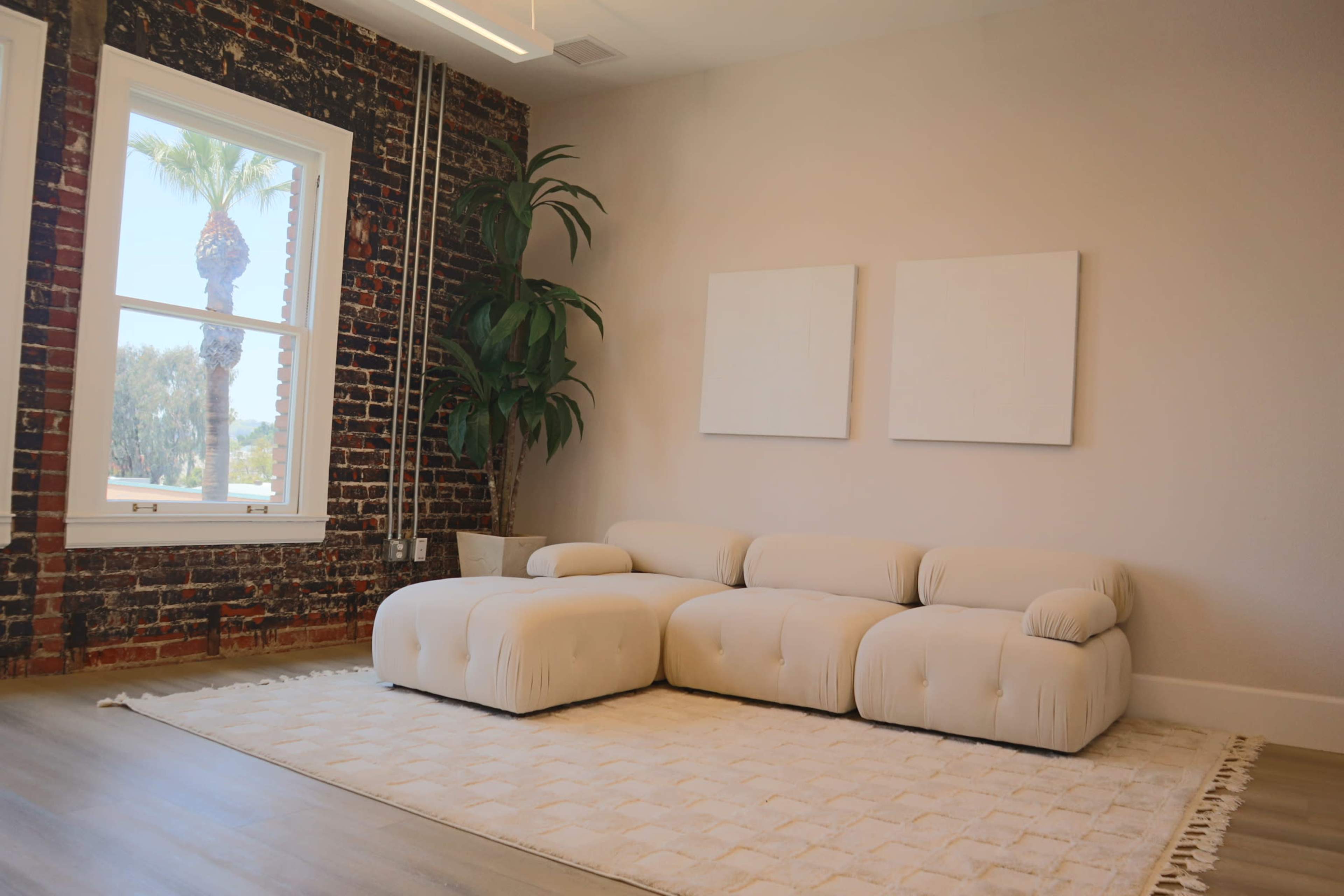 A modern living area featuring a light-colored sectional sofa, two blank white canvas frames on the wall, and a large window with a view of palm trees, contrasted against a red brick wall.