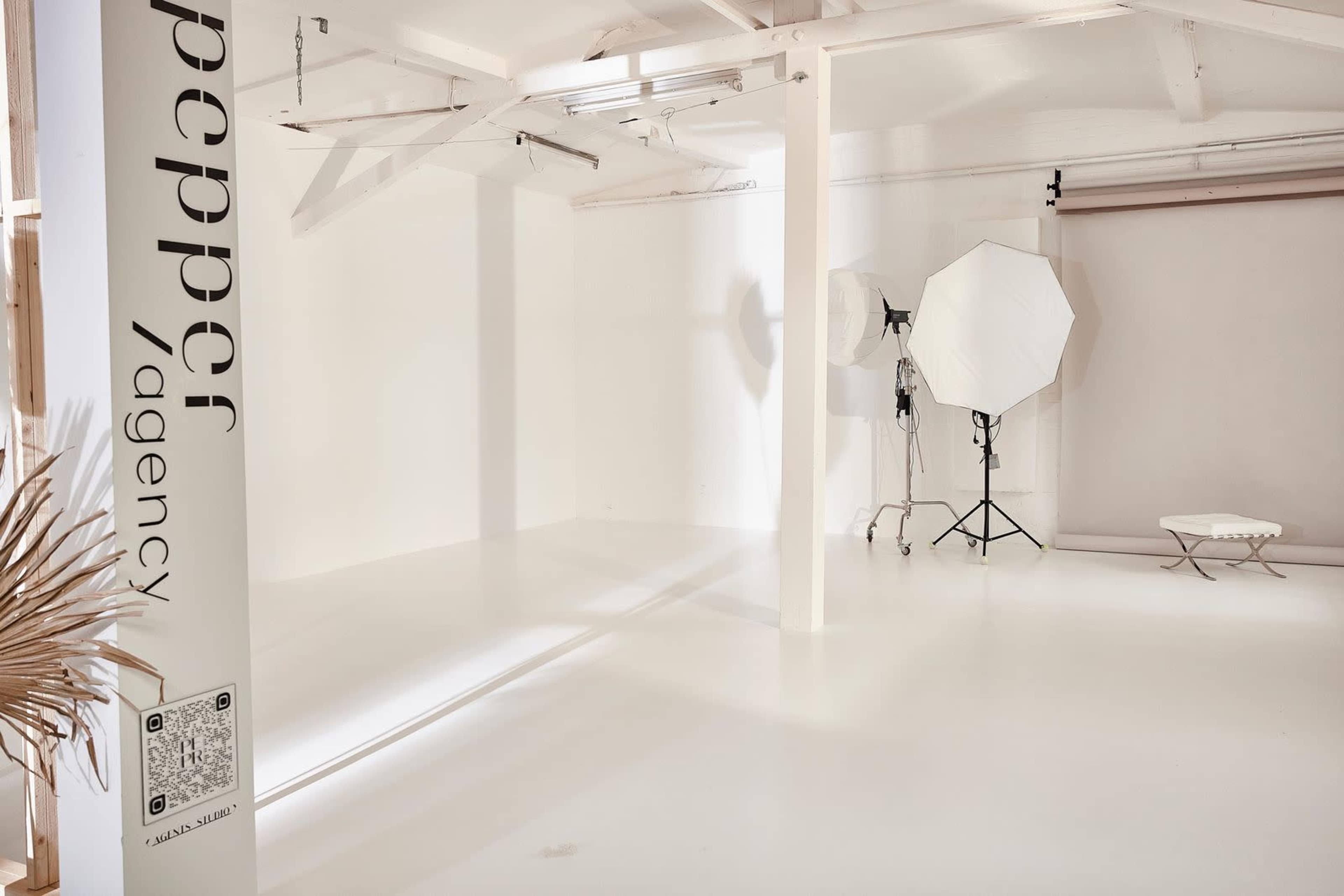 The image shows a minimalistic studio space with white walls, a backdrop setup, a large softbox on a stand, and a low table.