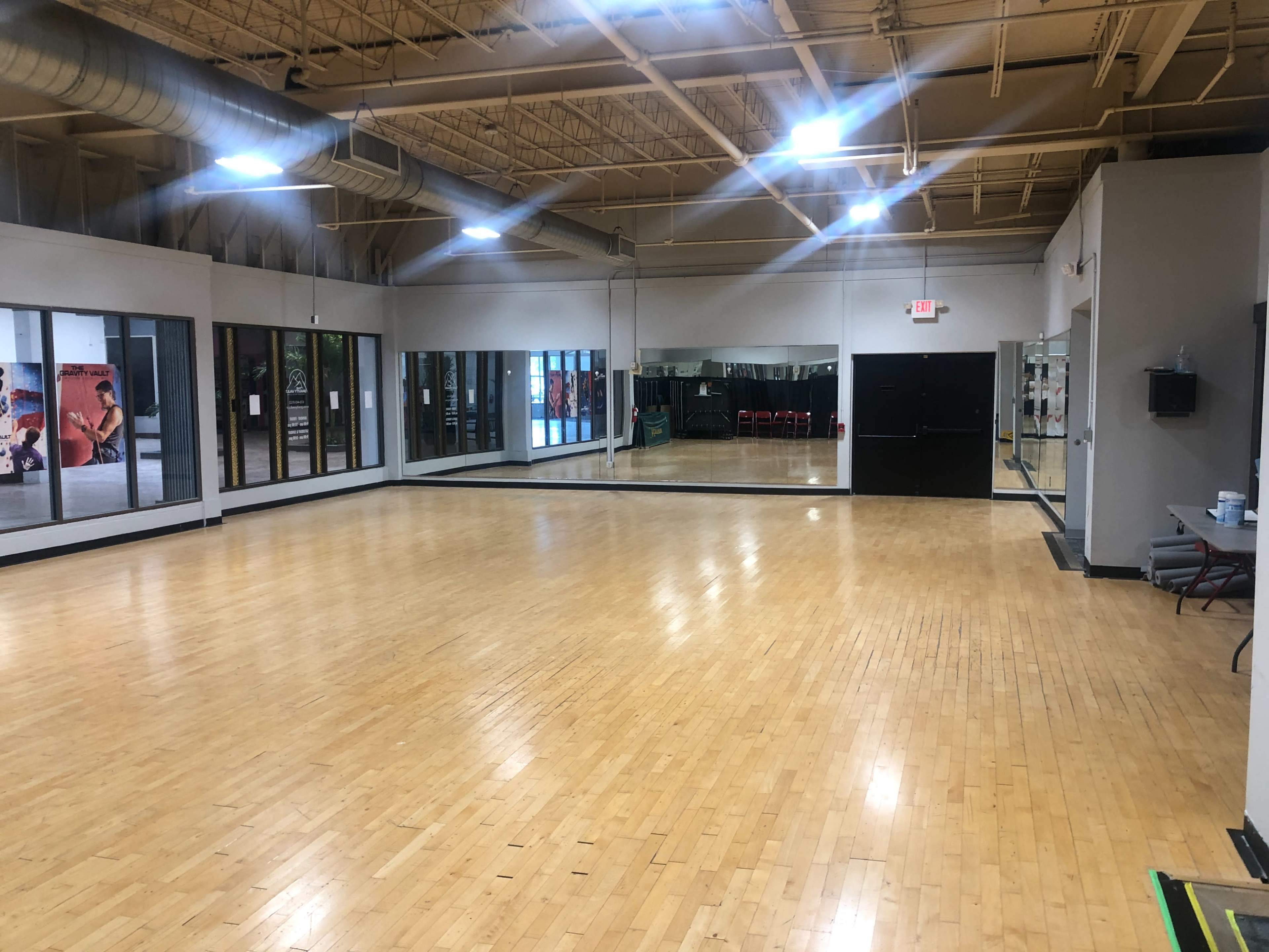 The image shows an empty indoor gym studio with wooden flooring and large windows that reflect the mirrored walls.