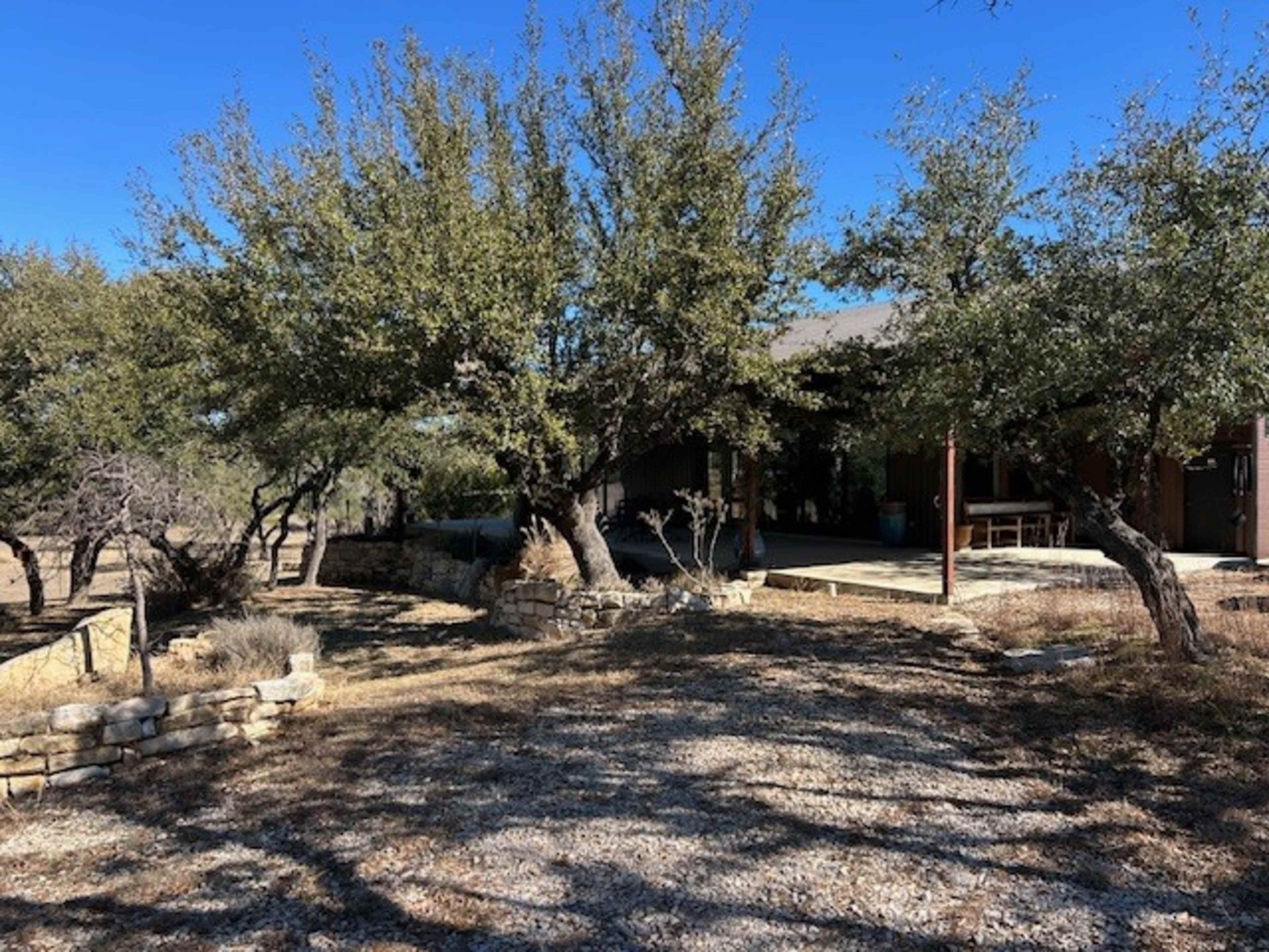The image shows a rustic house in a partly wooded area with several trees and a gravel path leading to the entrance.