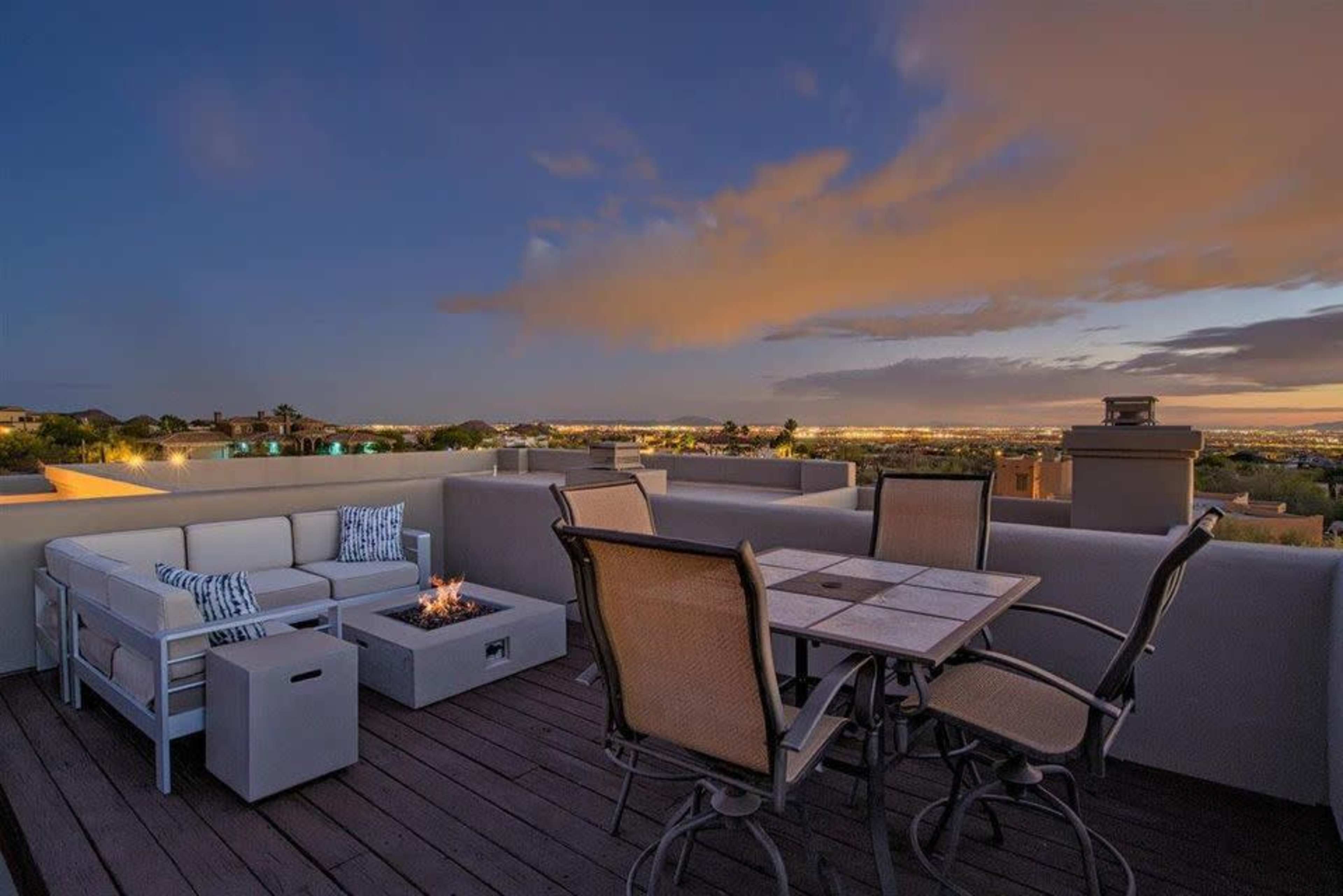 A rooftop patio with seating arrangements, a fire pit, and a view of the city skyline under a twilight sky.