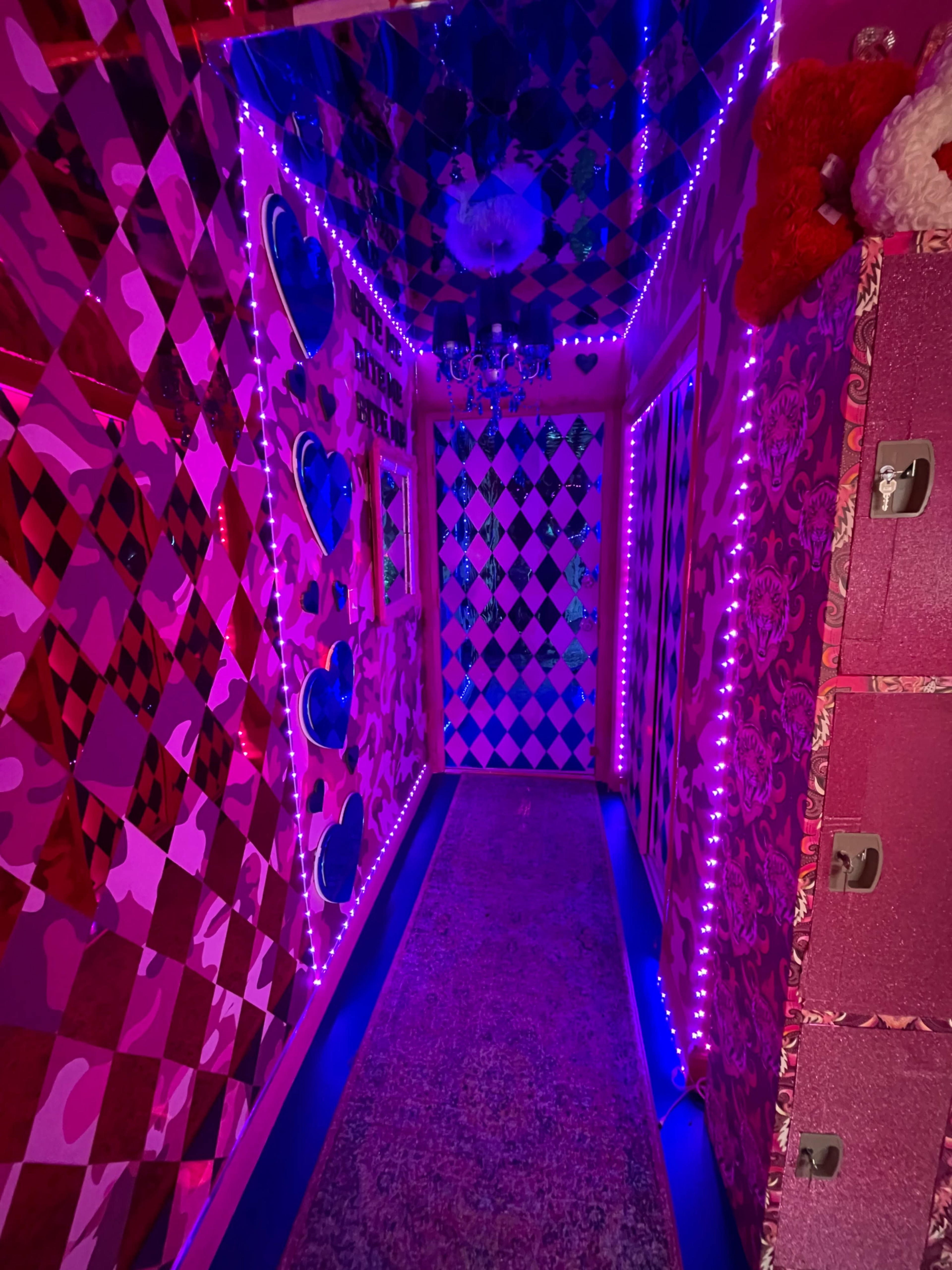 The scene depicts a brightly lit hallway decorated with pink and purple patterned walls, heart shapes, and neon lights, leading to a door at the end.