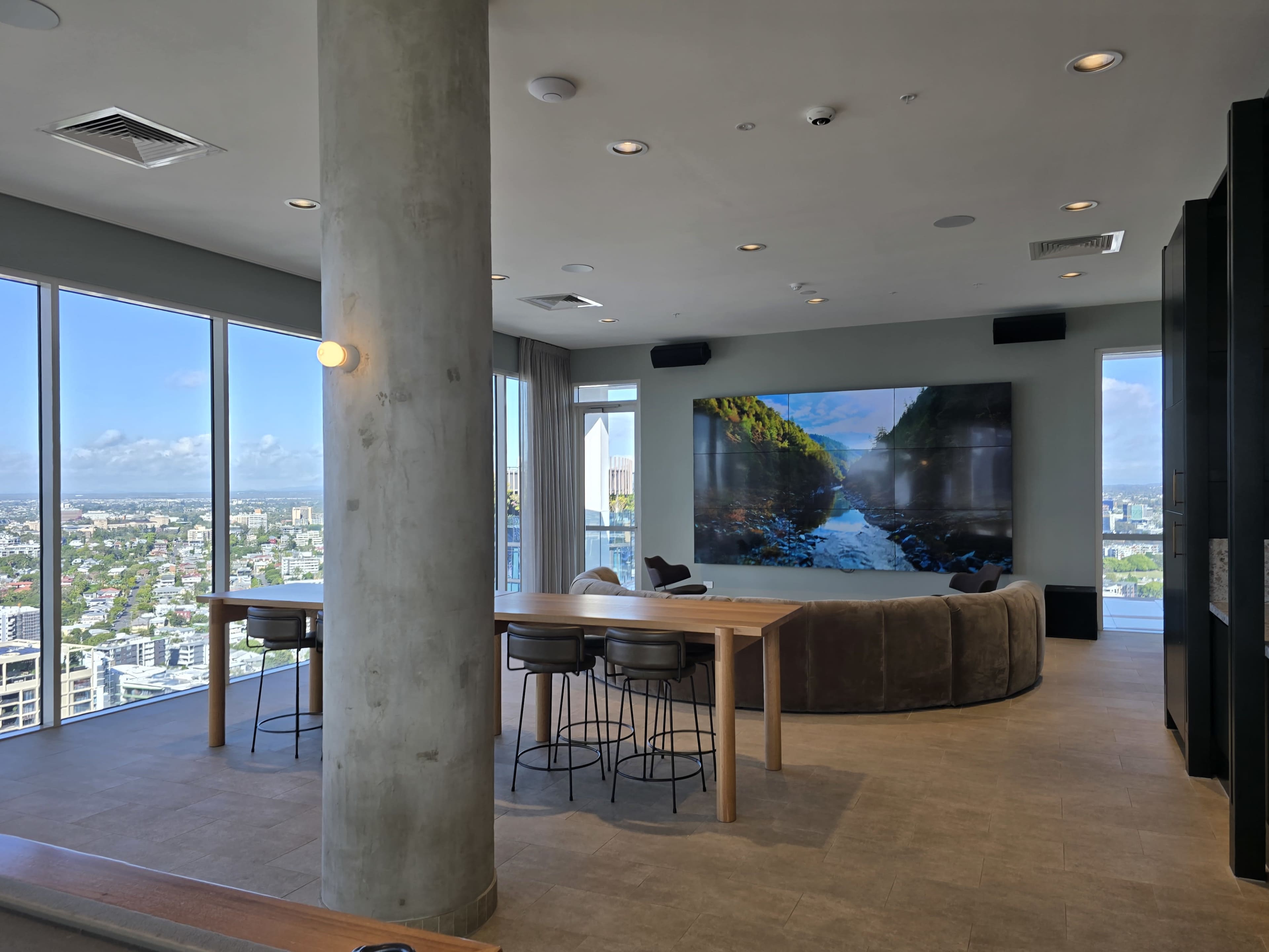 The image shows a modern lounge area with large windows offering a city view, a wooden dining table, and a large screen displaying a scenic landscape.