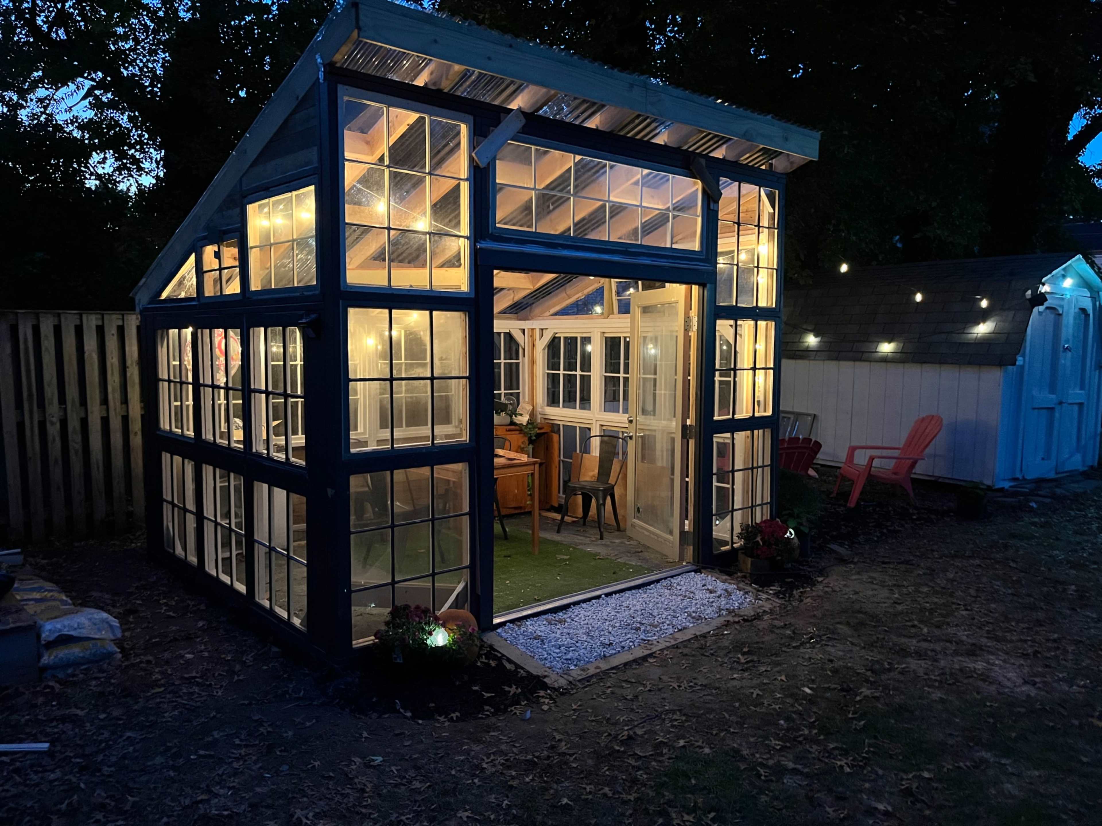 A well-lit greenhouse made of glass panels stands in a backyard, surrounded by outdoor seating and decorative lights.