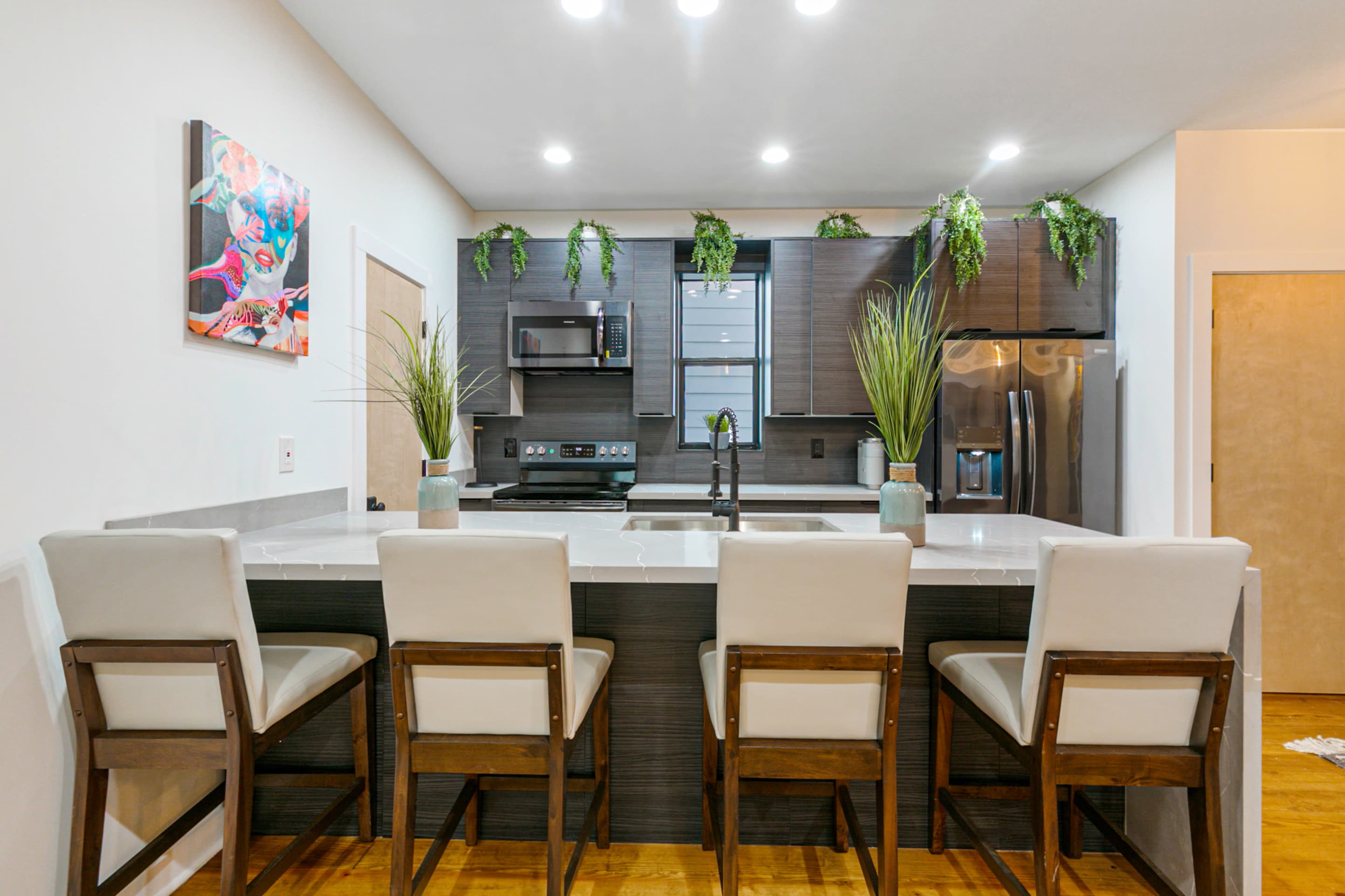 A modern kitchen features a counter with four upholstered stools, stainless steel appliances, and decorative greenery.