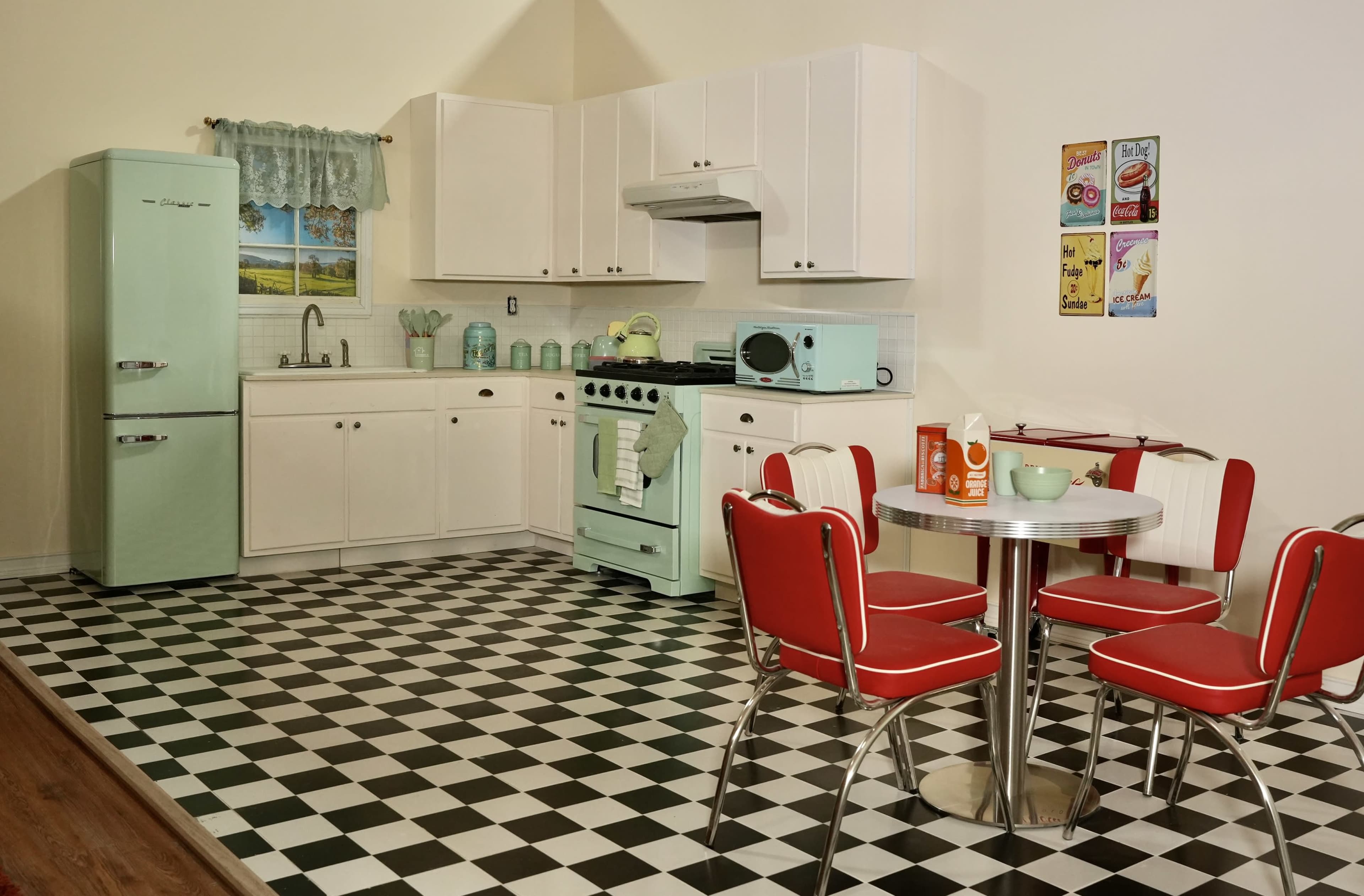 A retro-style kitchen features mint green appliances, white cabinets, a checkered black and white floor, and a red and silver dining set.