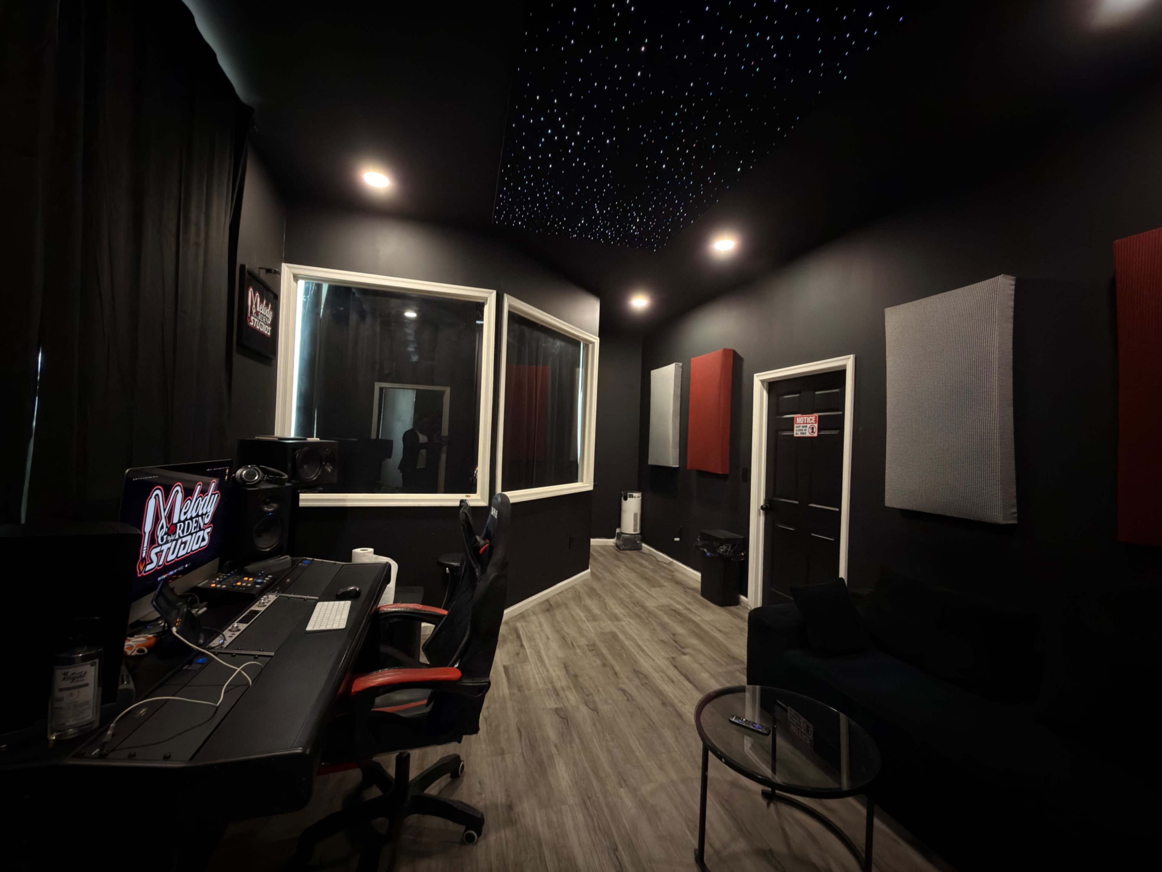 The image shows a recording studio with a soundproofed control room, a desk with audio equipment, and a comfortable seating area.