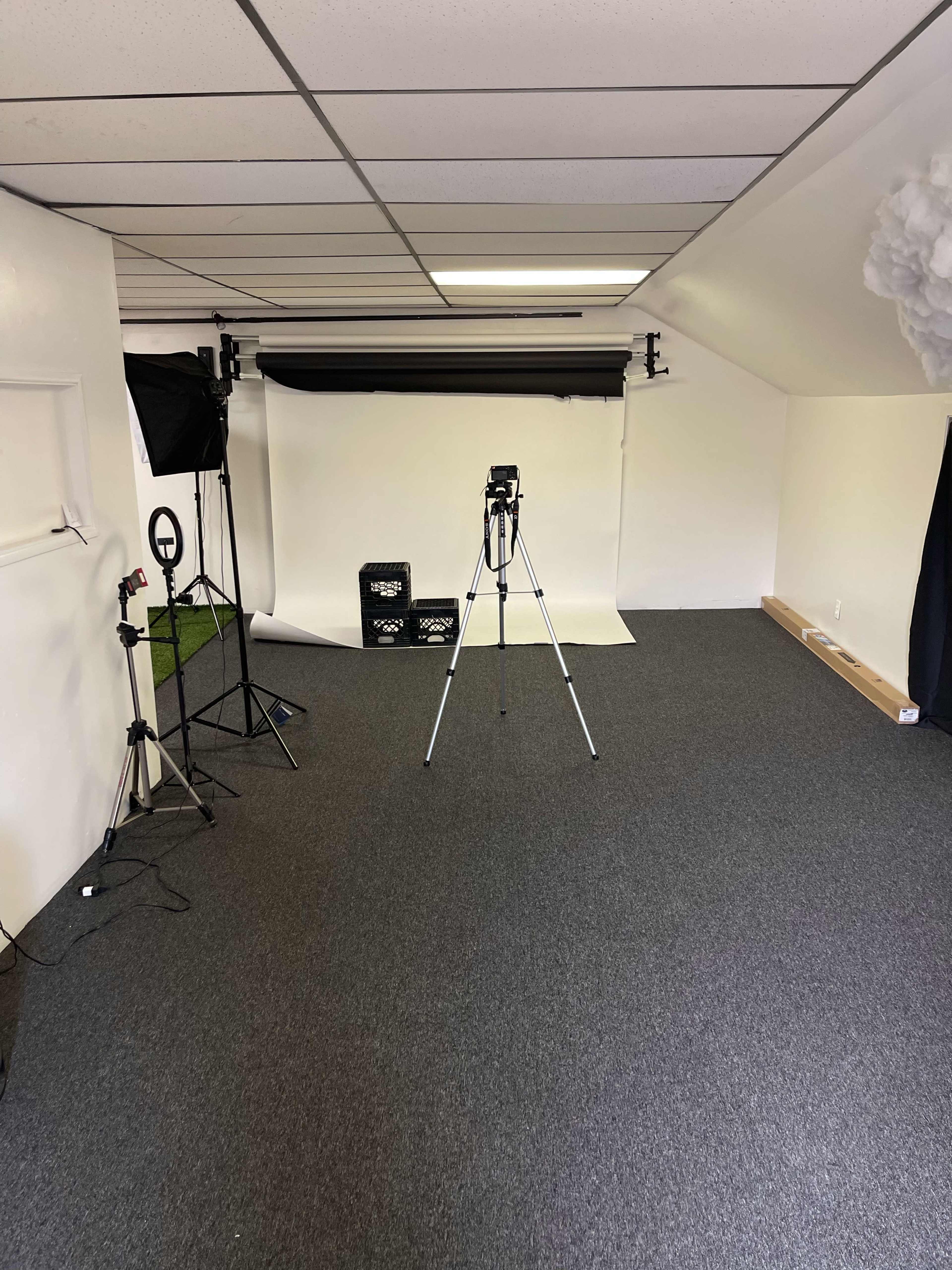 A blank studio space with a backdrop, a camera on a tripod, and lighting equipment set up in a room with gray carpeting.