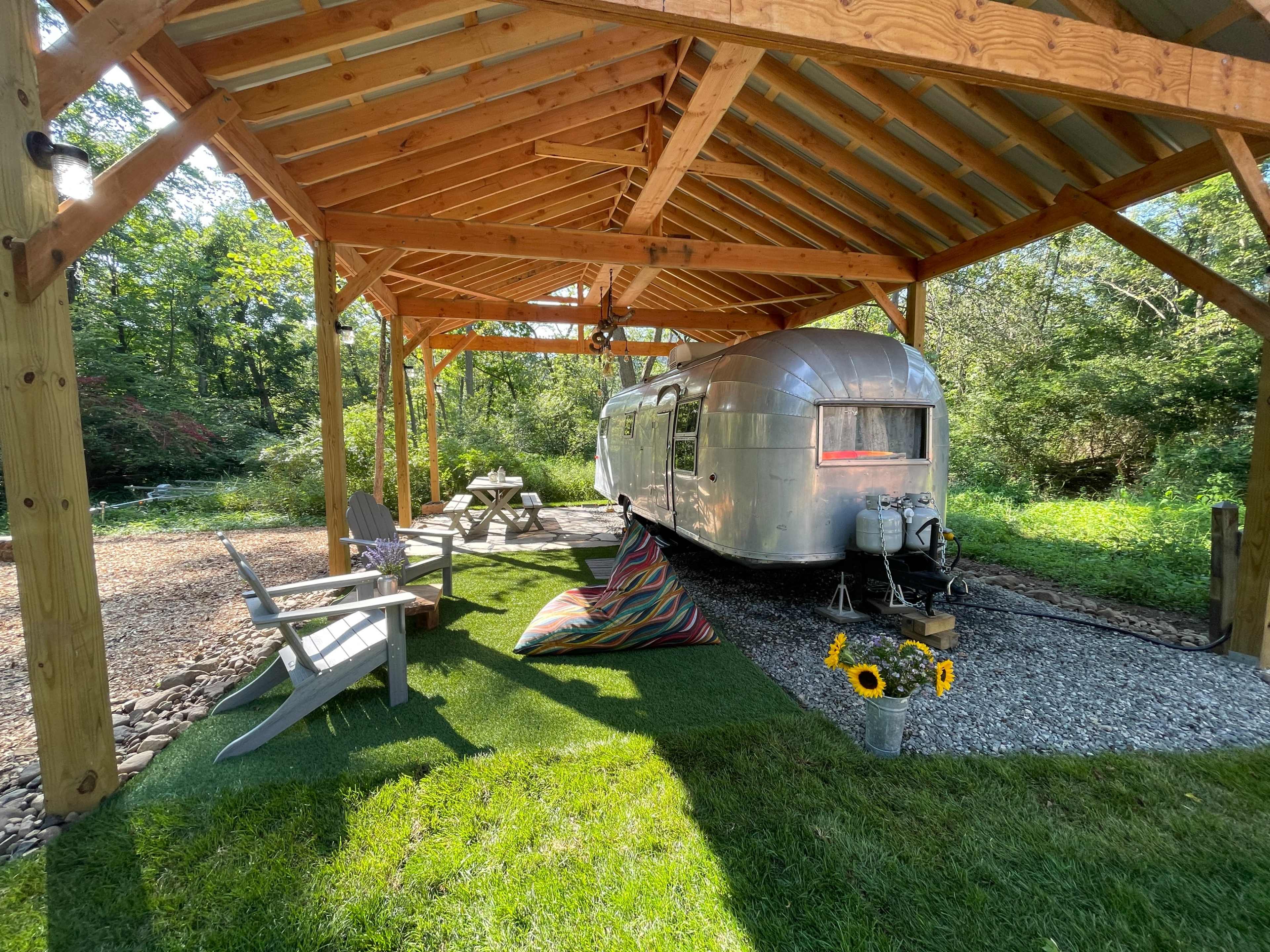 A vintage Airstream trailer is parked under a wooden shelter surrounded by a green lawn and decorative seating.