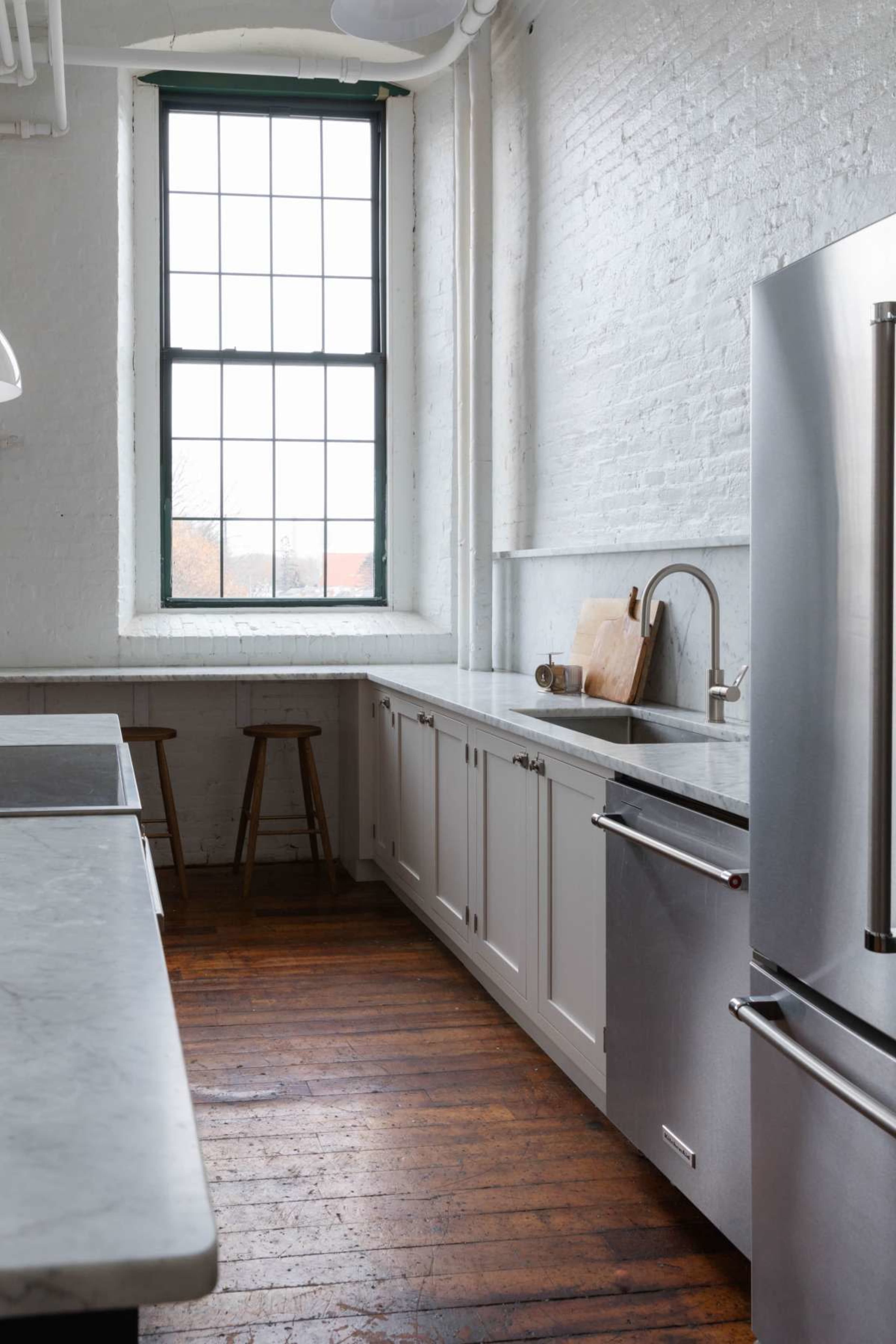 A modern kitchen features white brick walls, a large window, stainless steel appliances, and wooden flooring.