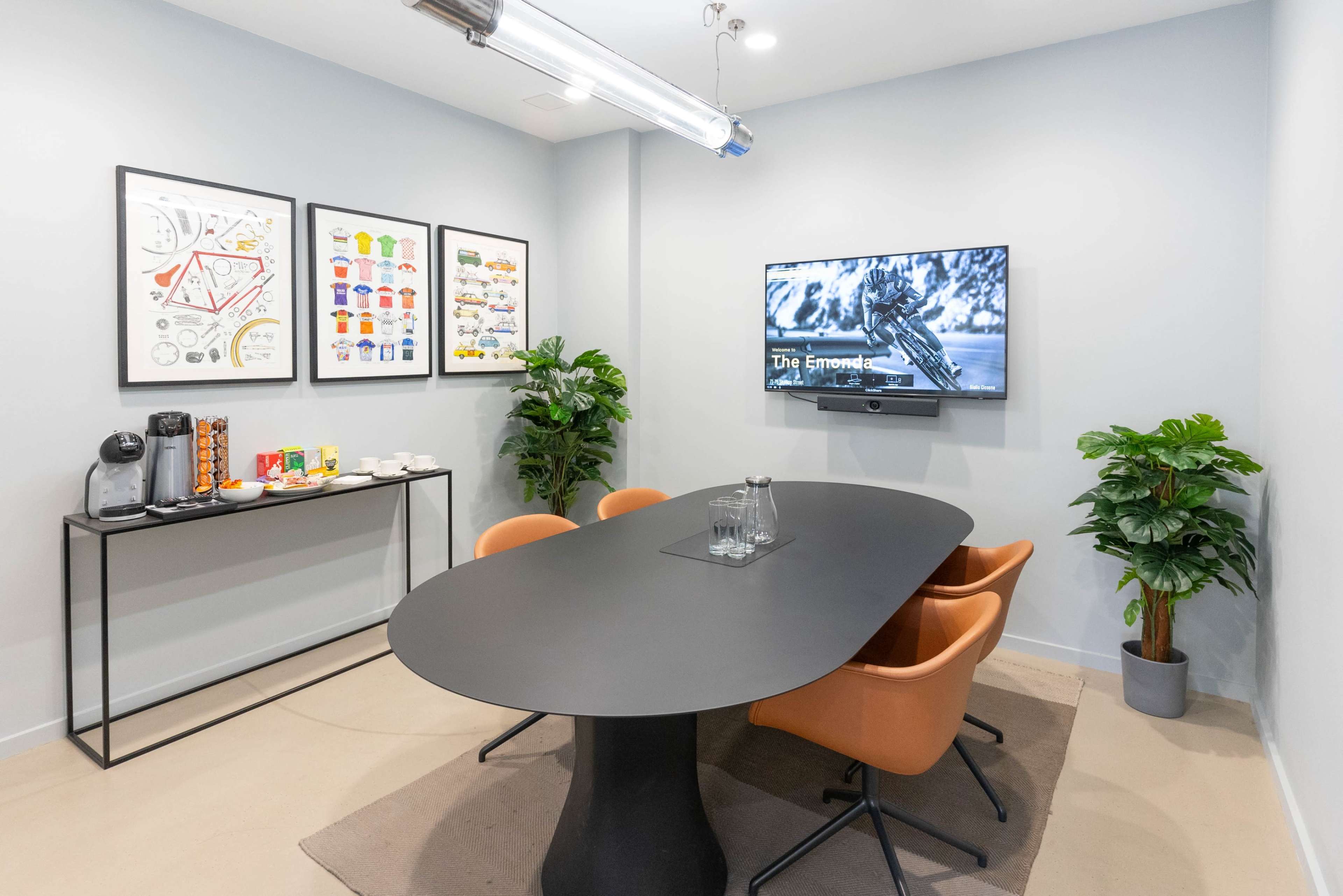 A modern meeting room features a black oval table surrounded by four orange chairs, a wall-mounted TV, plants, and framed art.