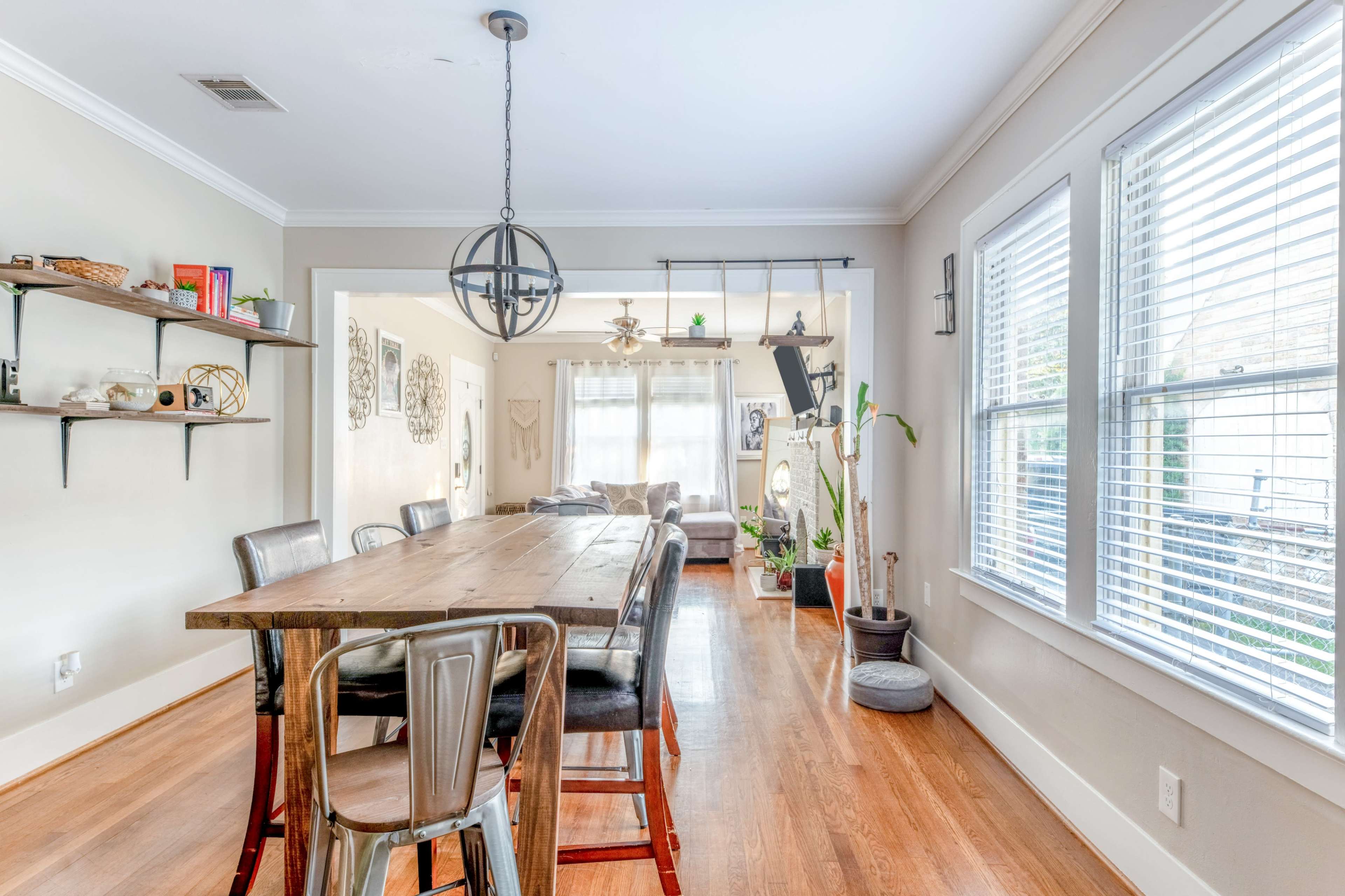 A wooden dining table with metal chairs is positioned in a bright, open living area featuring windows and decorative plants.