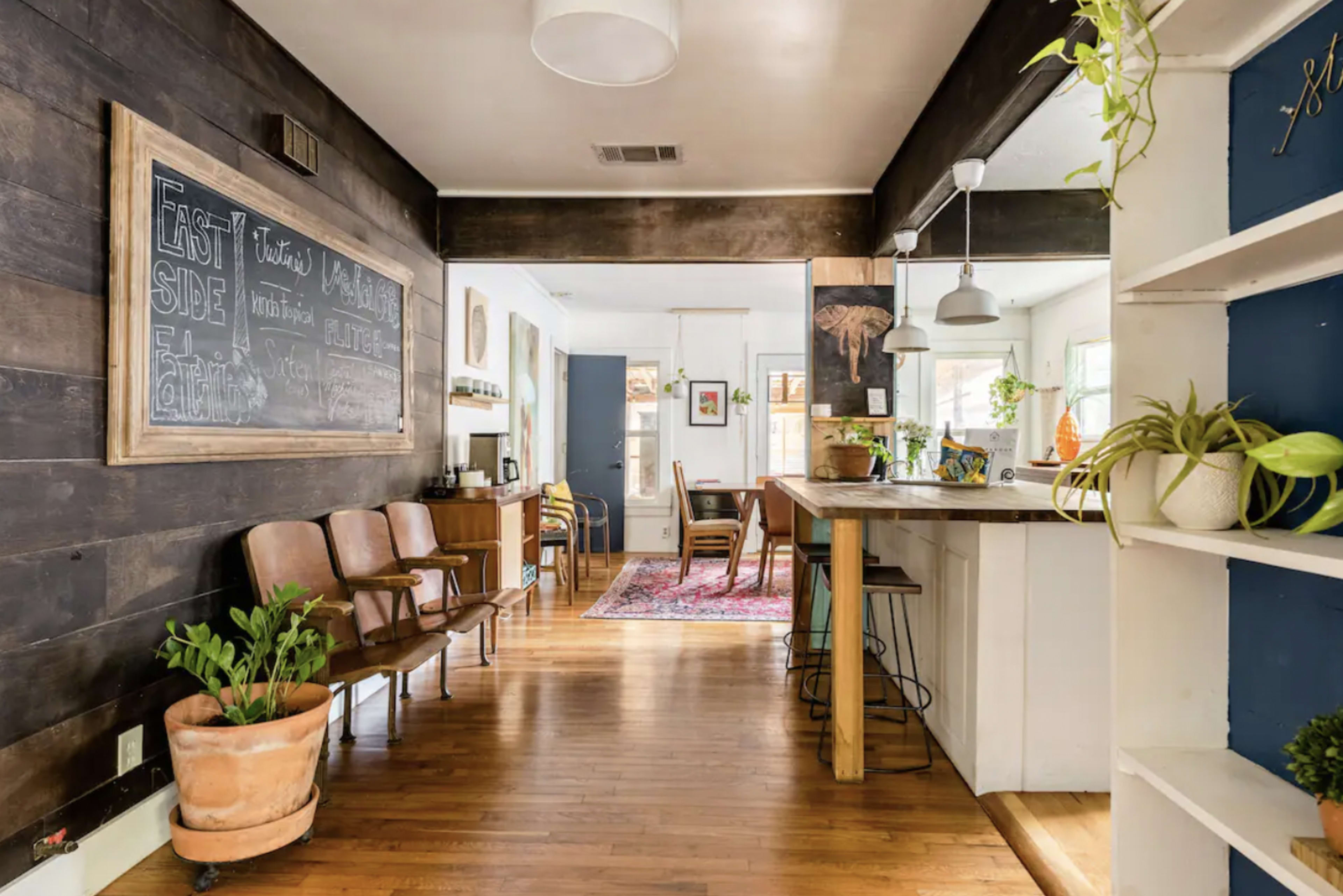 A spacious room with wooden flooring, featuring a hallway leading to a dining area and a kitchen, lined with plants and wooden furniture.
