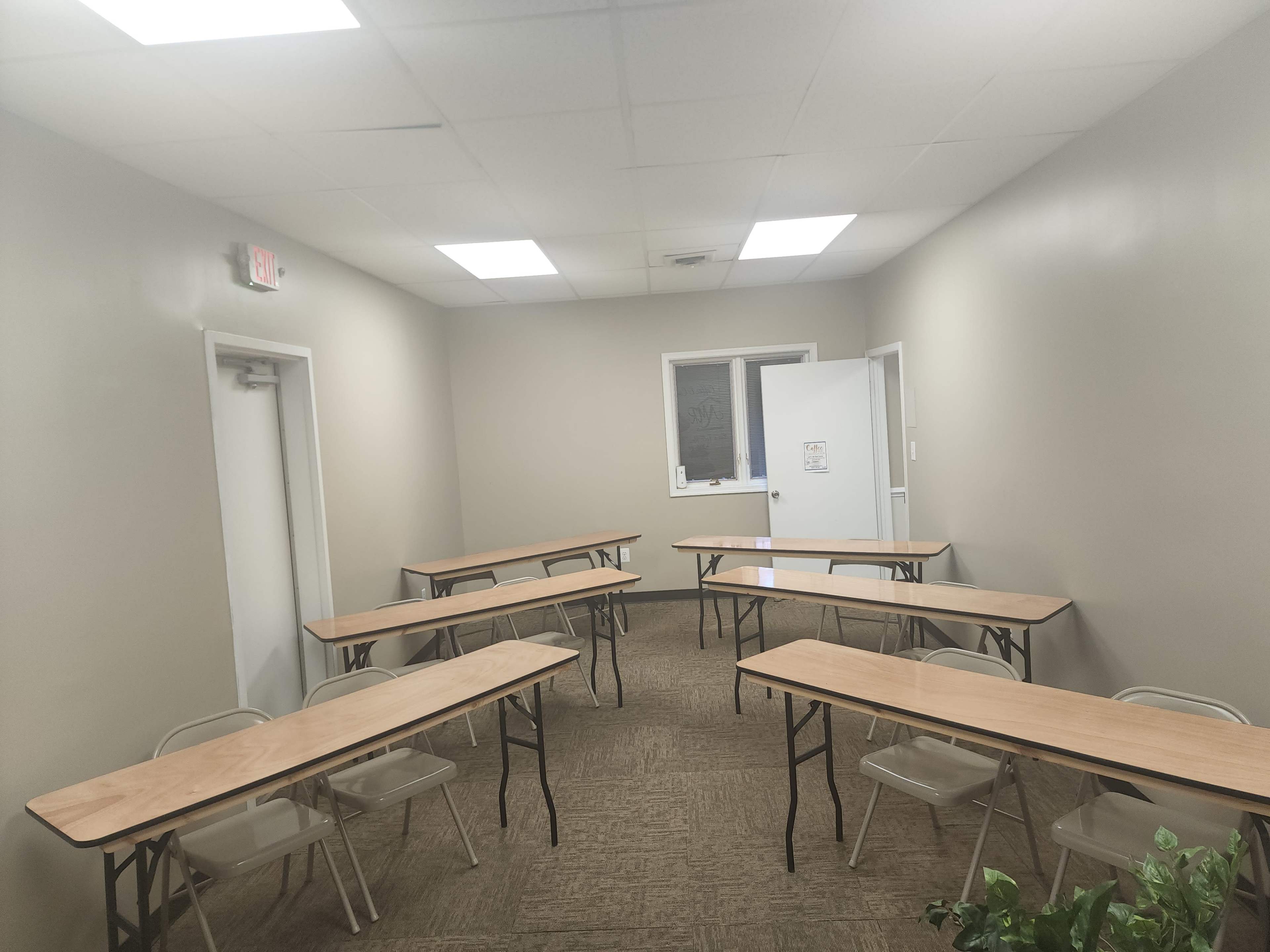 A small classroom setup with several wooden tables and folding chairs arranged in two rows in a plain, softly lit room.