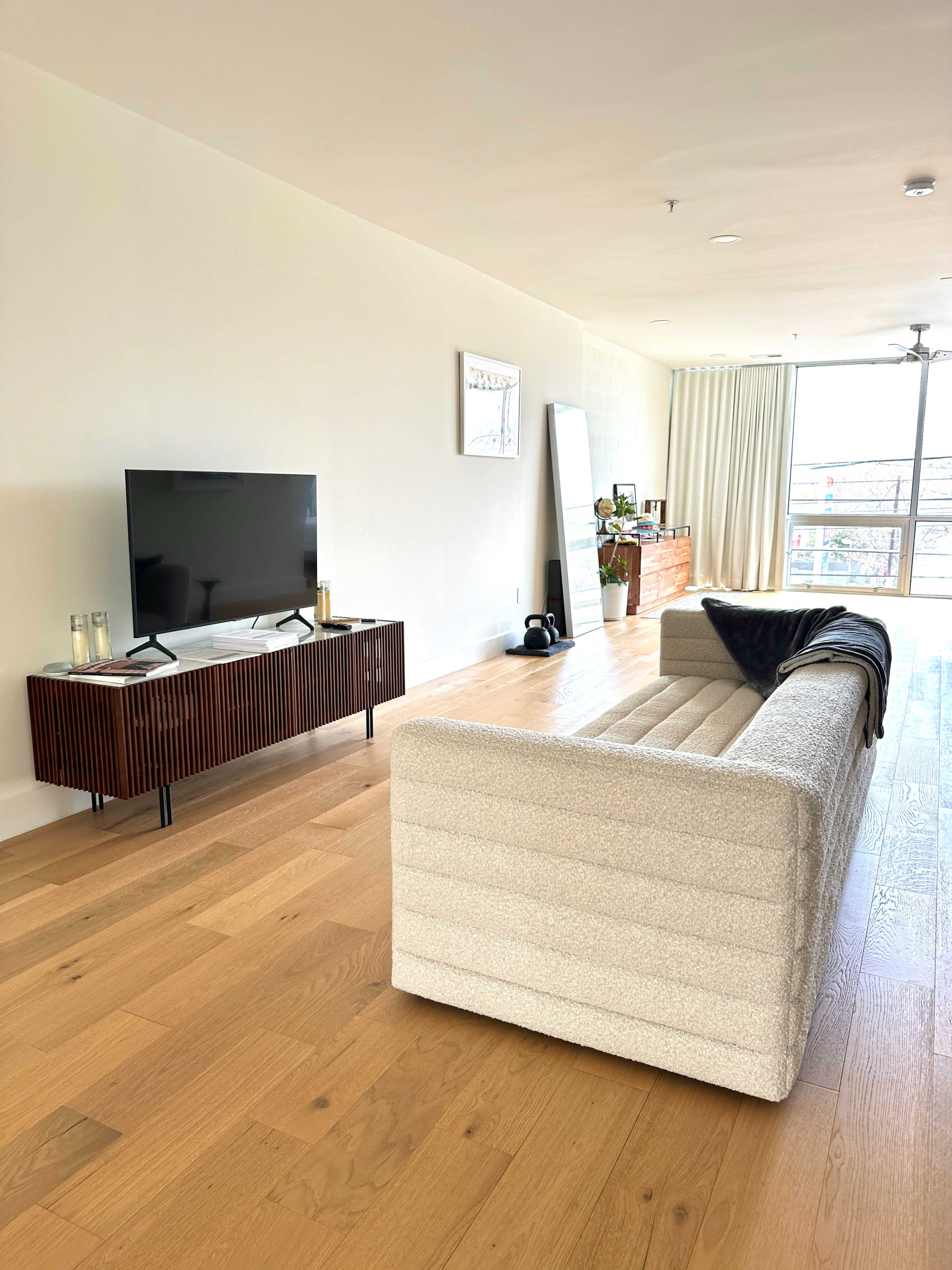 A modern living room features a light-colored sofa, a wooden TV stand with a television, and large windows with sheer curtains.